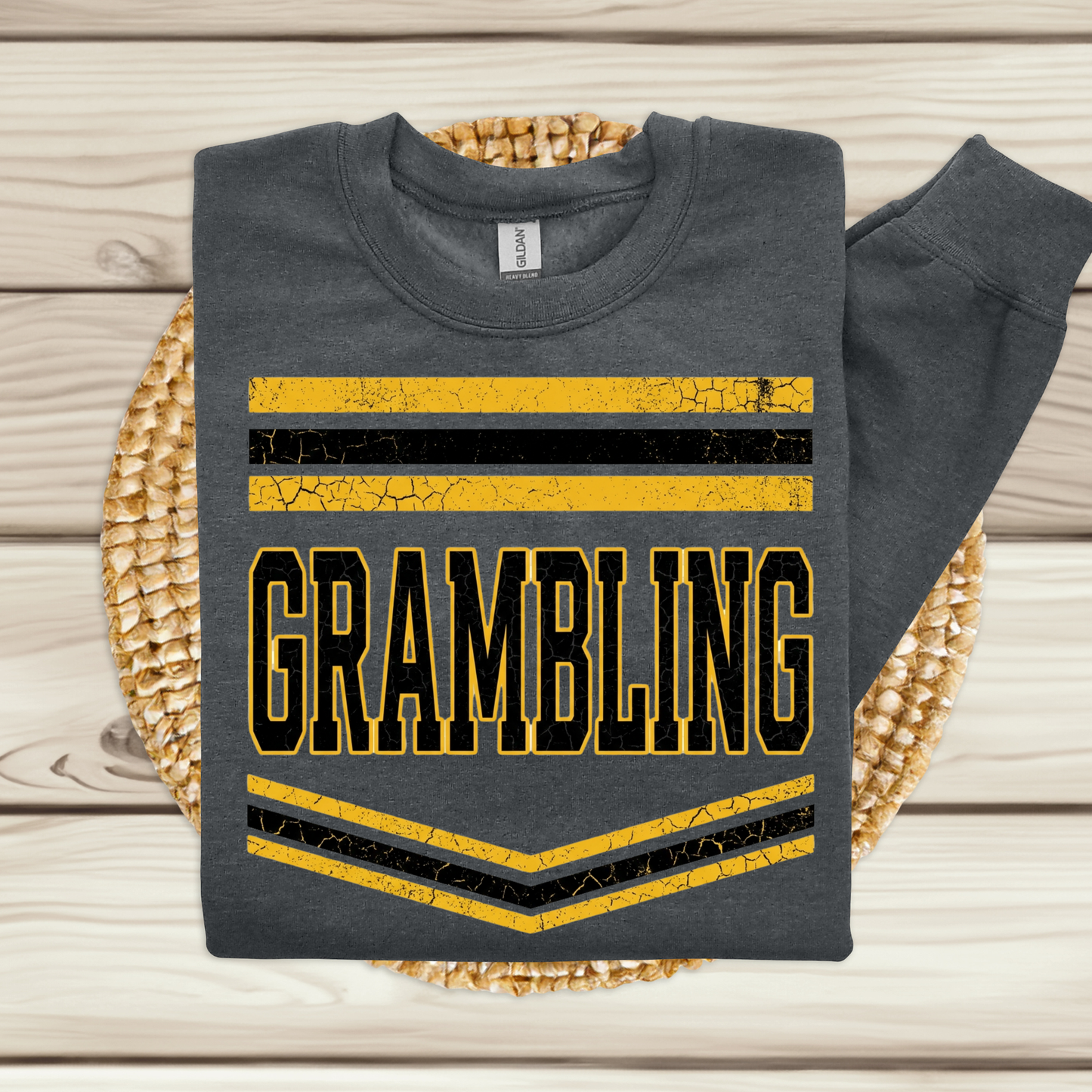 A dark gray sweatshirt laid flat on a woven round mat over a light wood plank surface. The sweatshirt has a distressed graphic with red and black horizontal stripes and the word 'PANTHERS' in large black letters outlined in red.