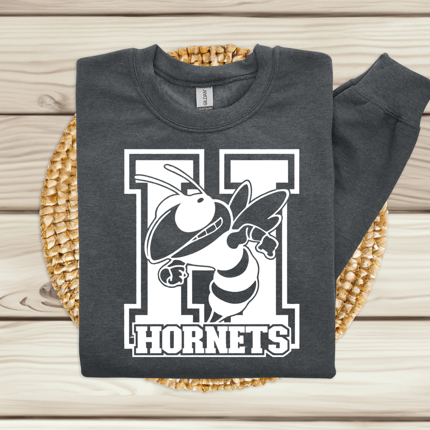 A dark charcoal gray sweatshirt with a white and black 'H Hornets' graphic, folded and displayed on a woven straw mat over a light wood plank background.
