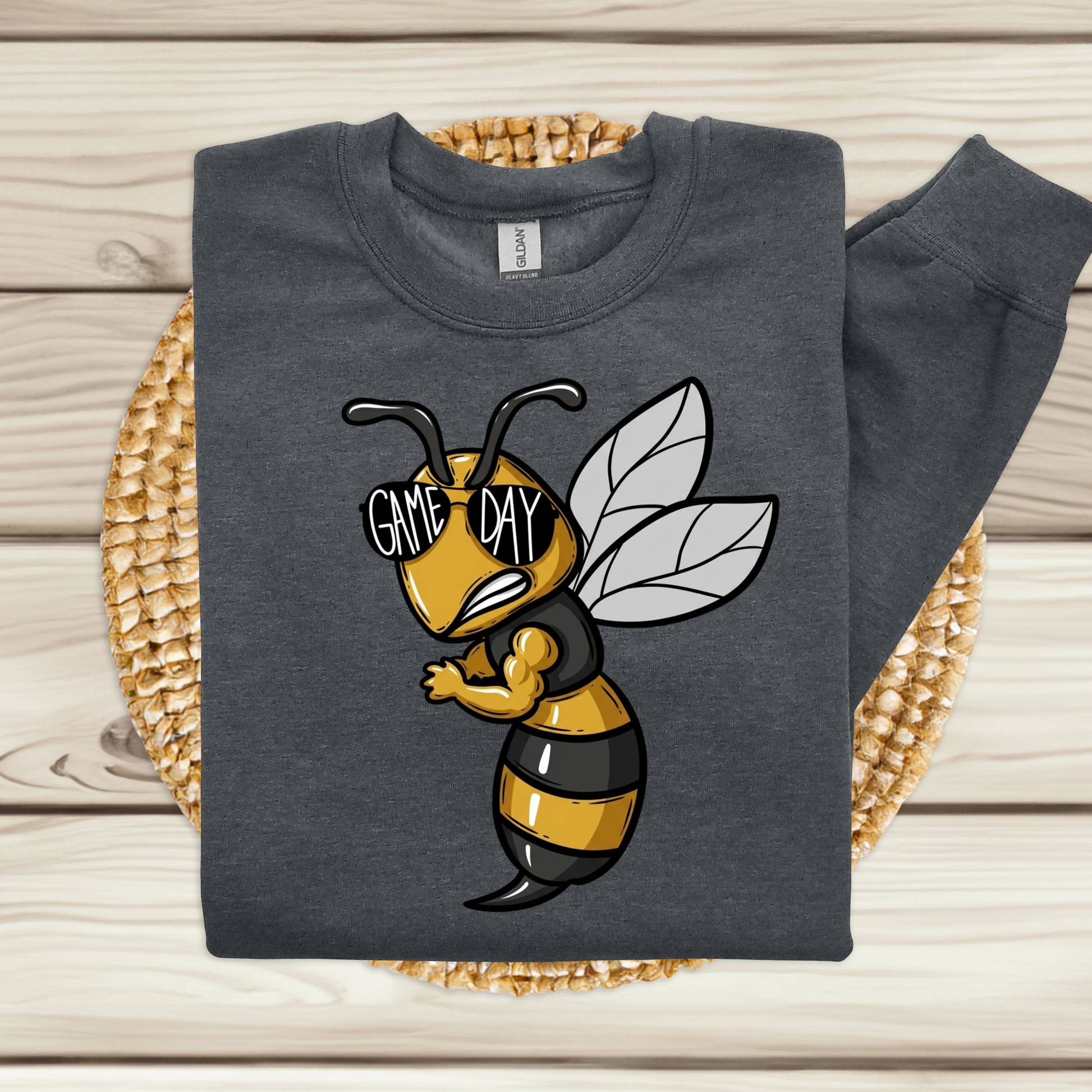 A dark gray sweatshirt laid flat on a woven round mat over a light wood plank surface. The sweatshirt has a large cartoon bee graphic on the front with the words “GAME DAY” across its eyes.