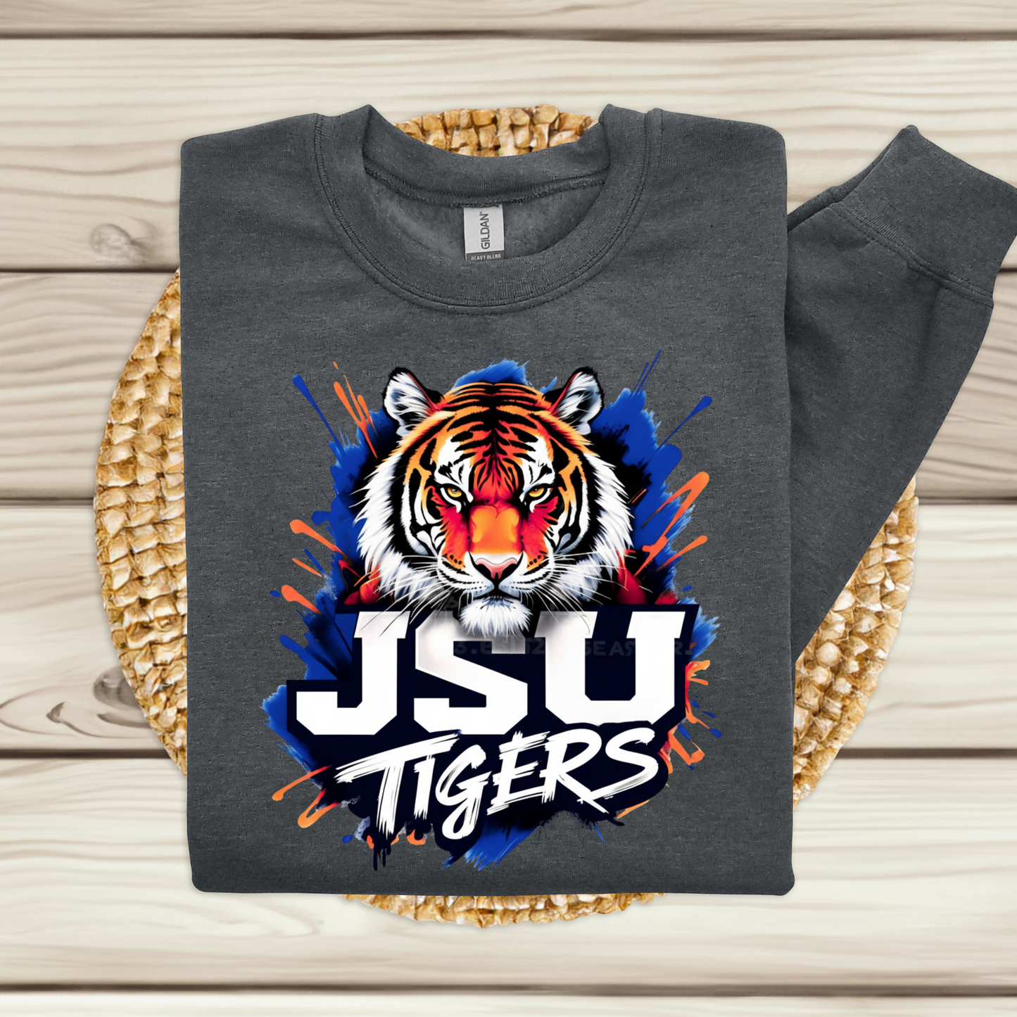 A dark gray sweatshirt laid flat on a woven round mat over light wood planks. The sweatshirt has a large front graphic of a tiger head with blue, orange, white, and black paint-splatter styling, plus the text 'JSU TIGERS' in large white letters with black outline.