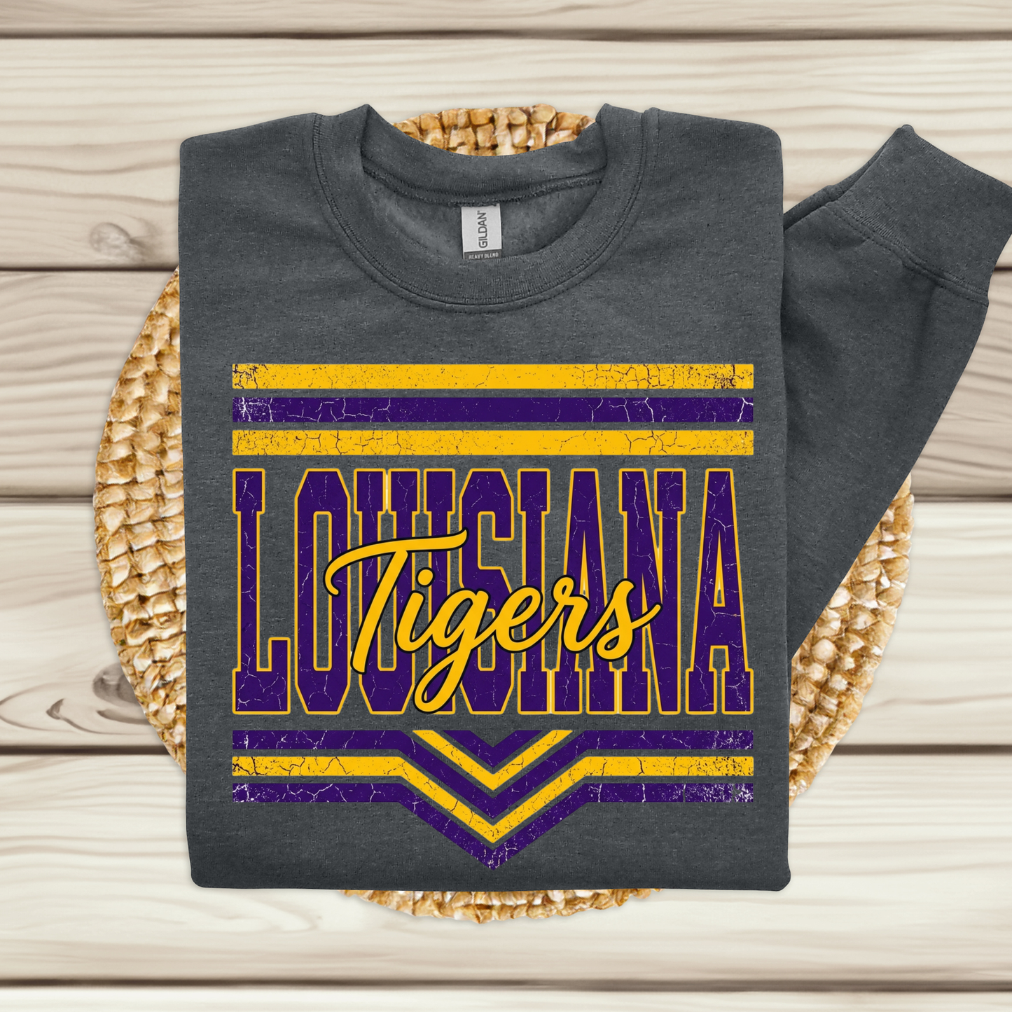 A dark gray sweatshirt with a distressed purple and yellow 'Louisiana Tigers' LSU graphic, folded and displayed on a woven placemat over a light wood background.