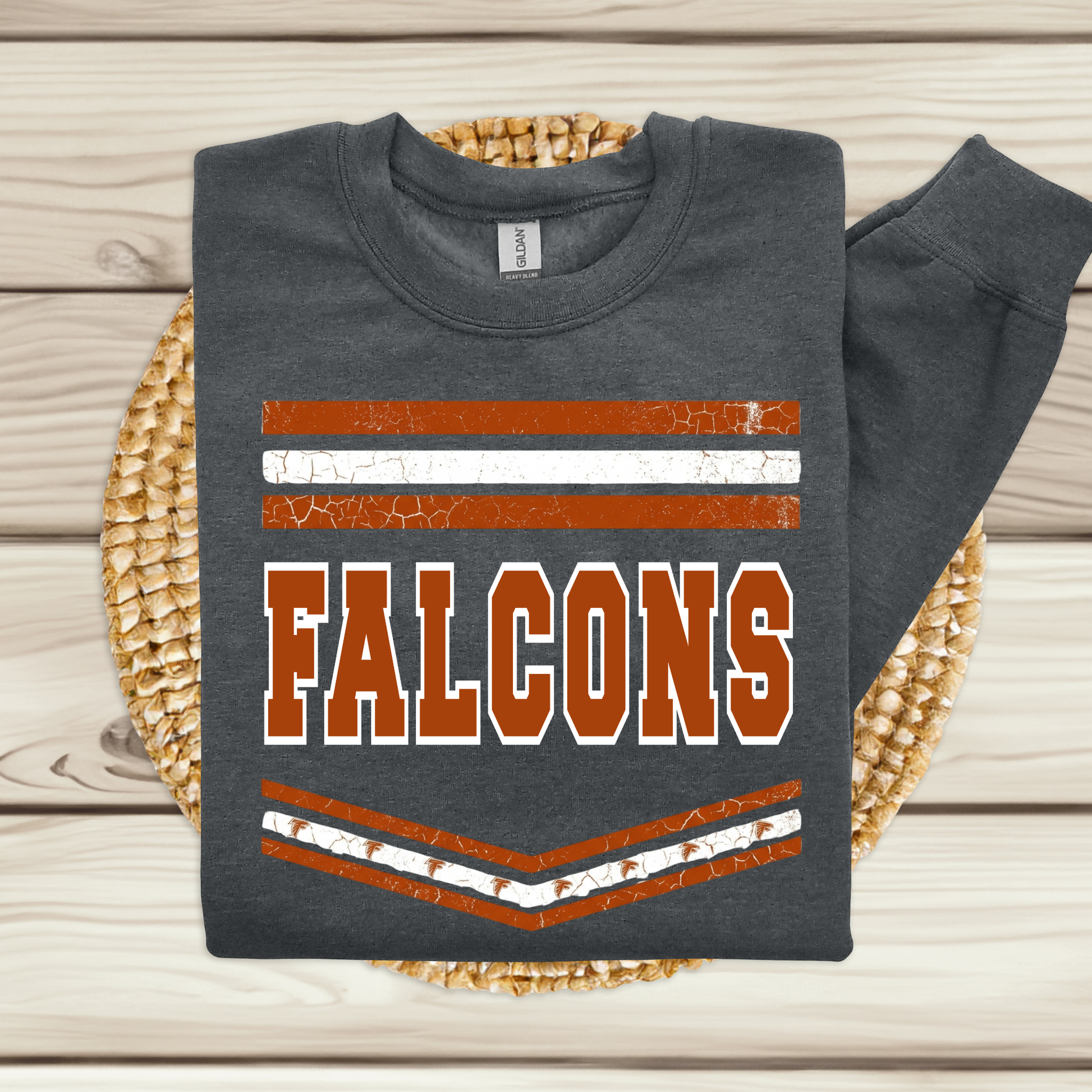 A dark gray sweatshirt with a distressed orange and white 'FALCONS' graphic, folded and displayed on a woven placemat over a light wood background.