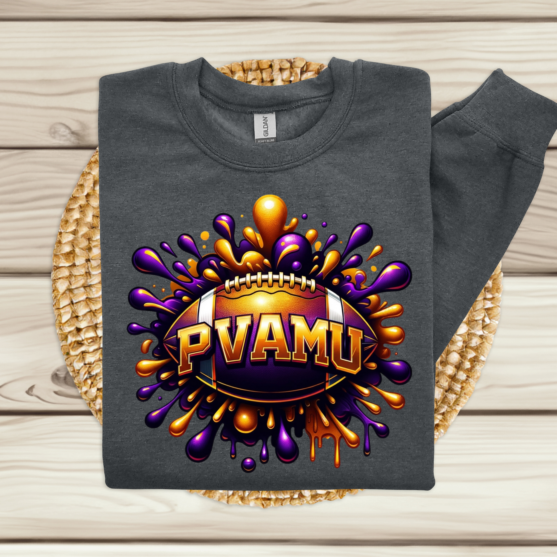A dark gray sweatshirt laid flat on a woven round mat over a light wood plank surface. The sweatshirt has a large front graphic of a football surrounded by purple and gold paint splatters, with the text 'PVAMU' across the center.