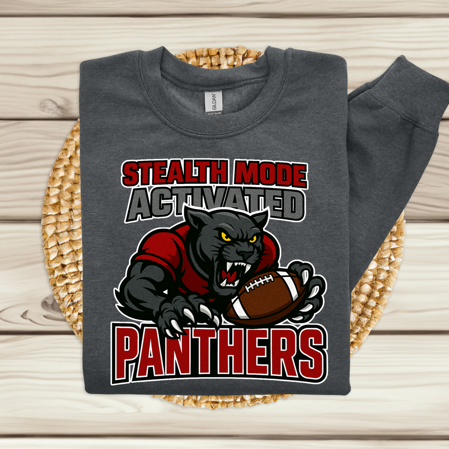 A dark gray long-sleeve t-shirt with a 'Stealth Mode Activated Panthers' graphic, displayed flat on a woven straw mat over a light wood plank background.