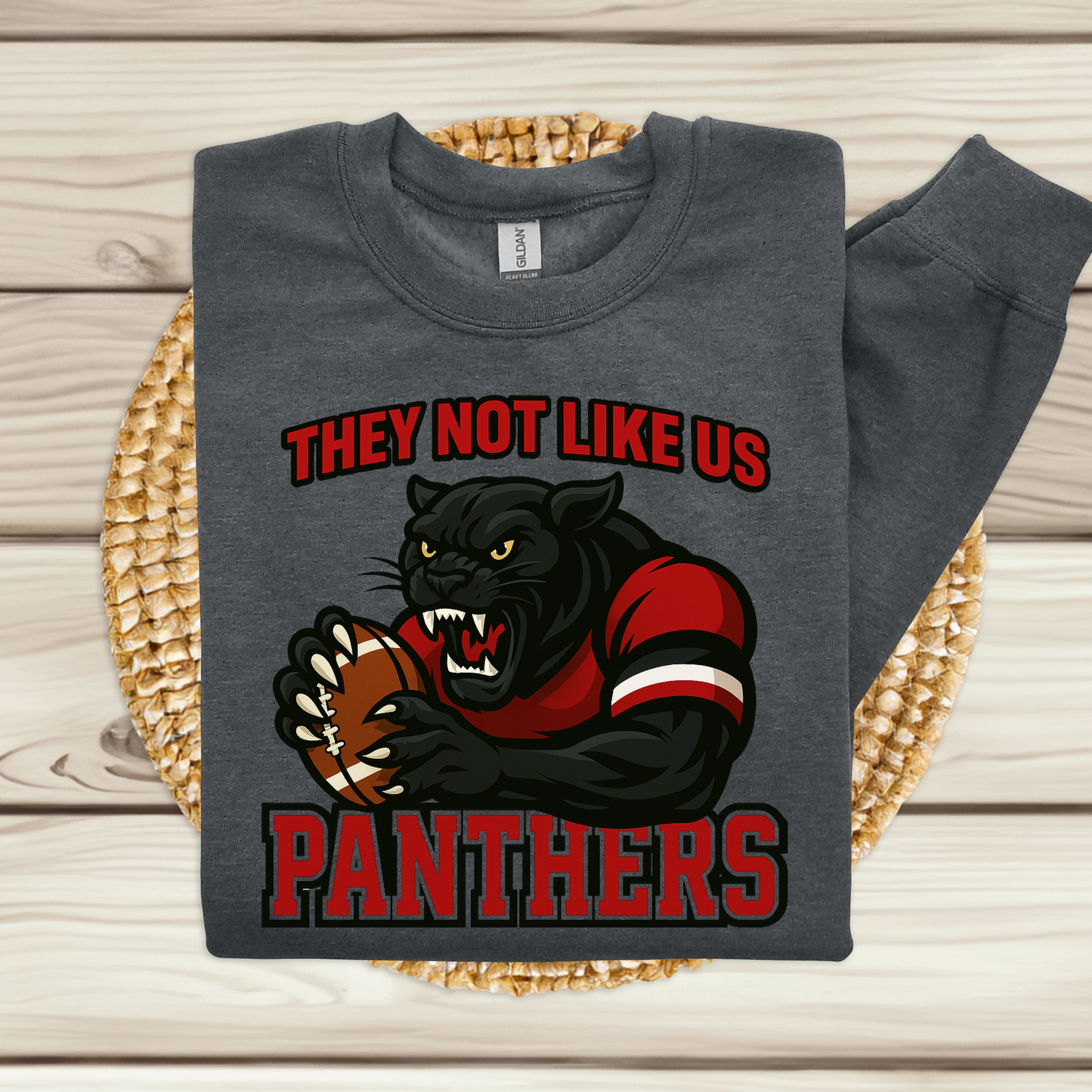 A dark gray sweatshirt laid flat on a woven round mat over a light wood plank background. The sweatshirt has a large graphic of a black panther holding a football, with red and black text reading 'THEY NOT LIKE US PANTHERS'.