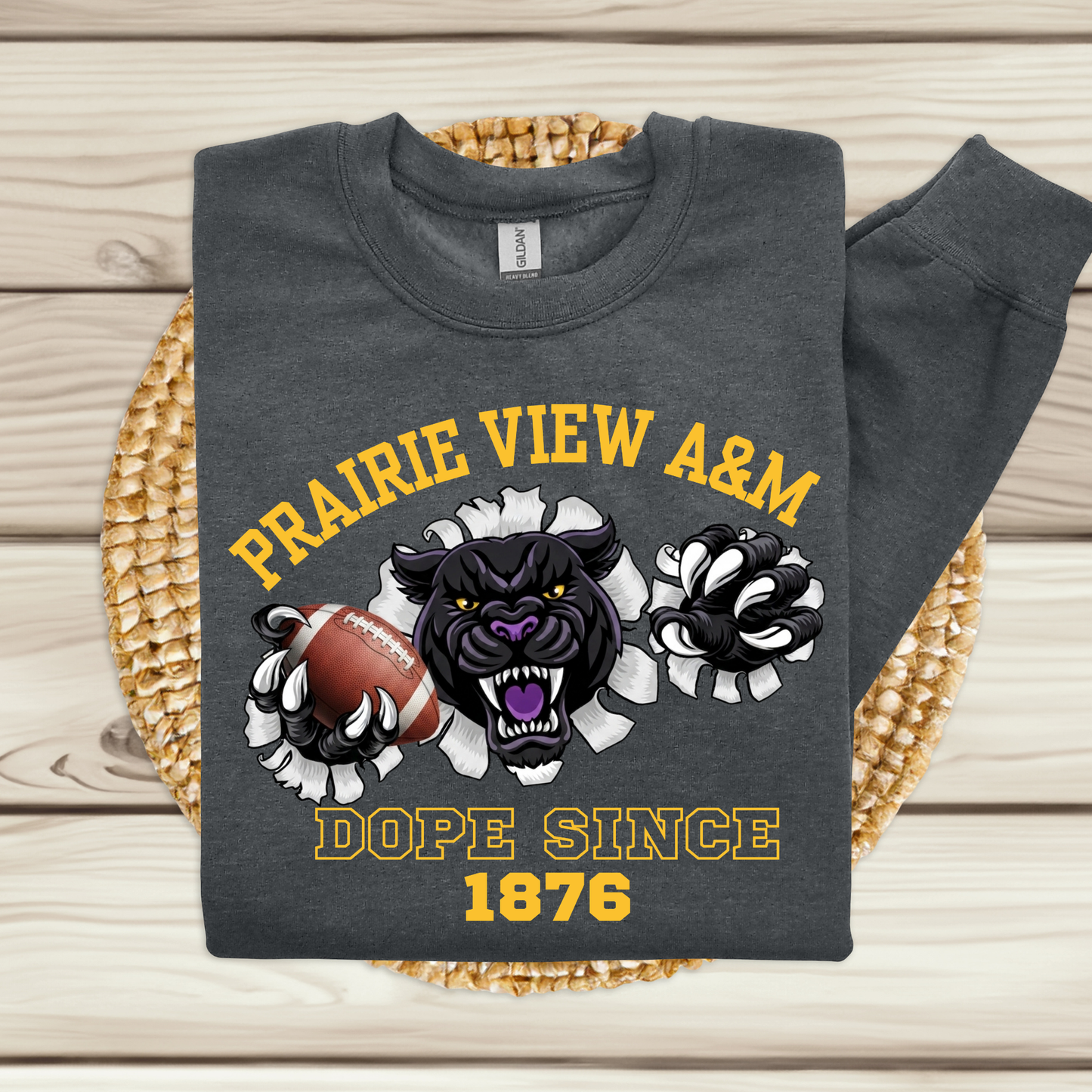 A dark gray sweatshirt laid flat on a woven round mat over a light wood surface. The sweatshirt has a large graphic print featuring the text 'PRAIRIE VIEW A&M' and 'DOPE SINCE 1876' in yellow, a black panther mascot, a football, and white flower motifs.