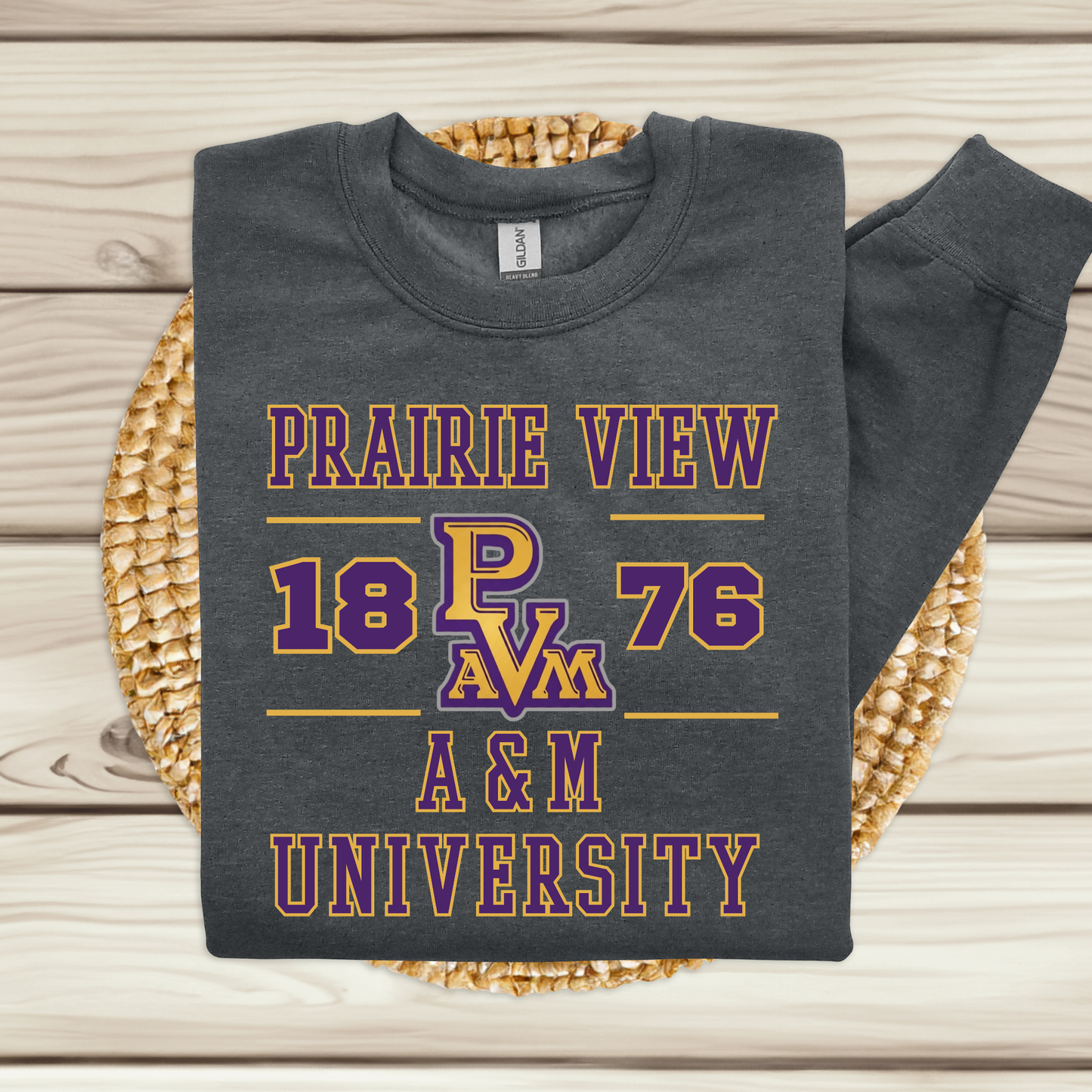 A dark charcoal gray sweatshirt with a purple and gold graphic reading 'PRAIRIE VIEW 18 PAVM 76 A & M UNIVERSITY,' folded and displayed on a woven round mat over a light wood plank background.