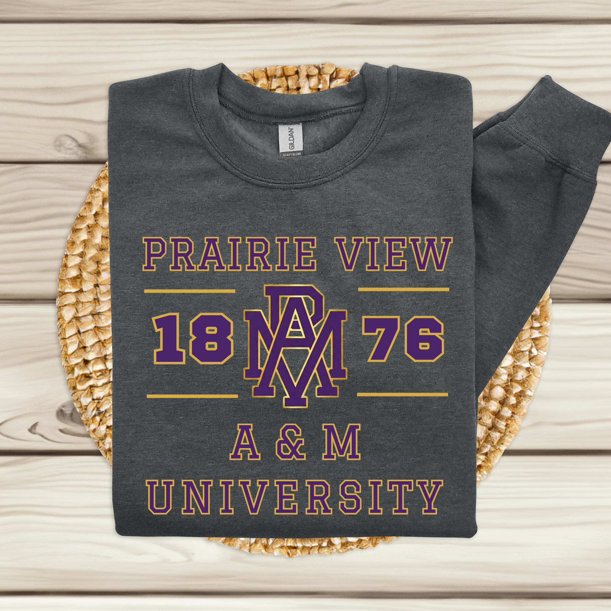 A dark charcoal gray sweatshirt with a purple and pink graphic print reading 'PRAIRIE VIEW 1876 A & M UNIVERSITY' and a large 'AM' monogram, laid flat on a woven round mat over light wood planks.