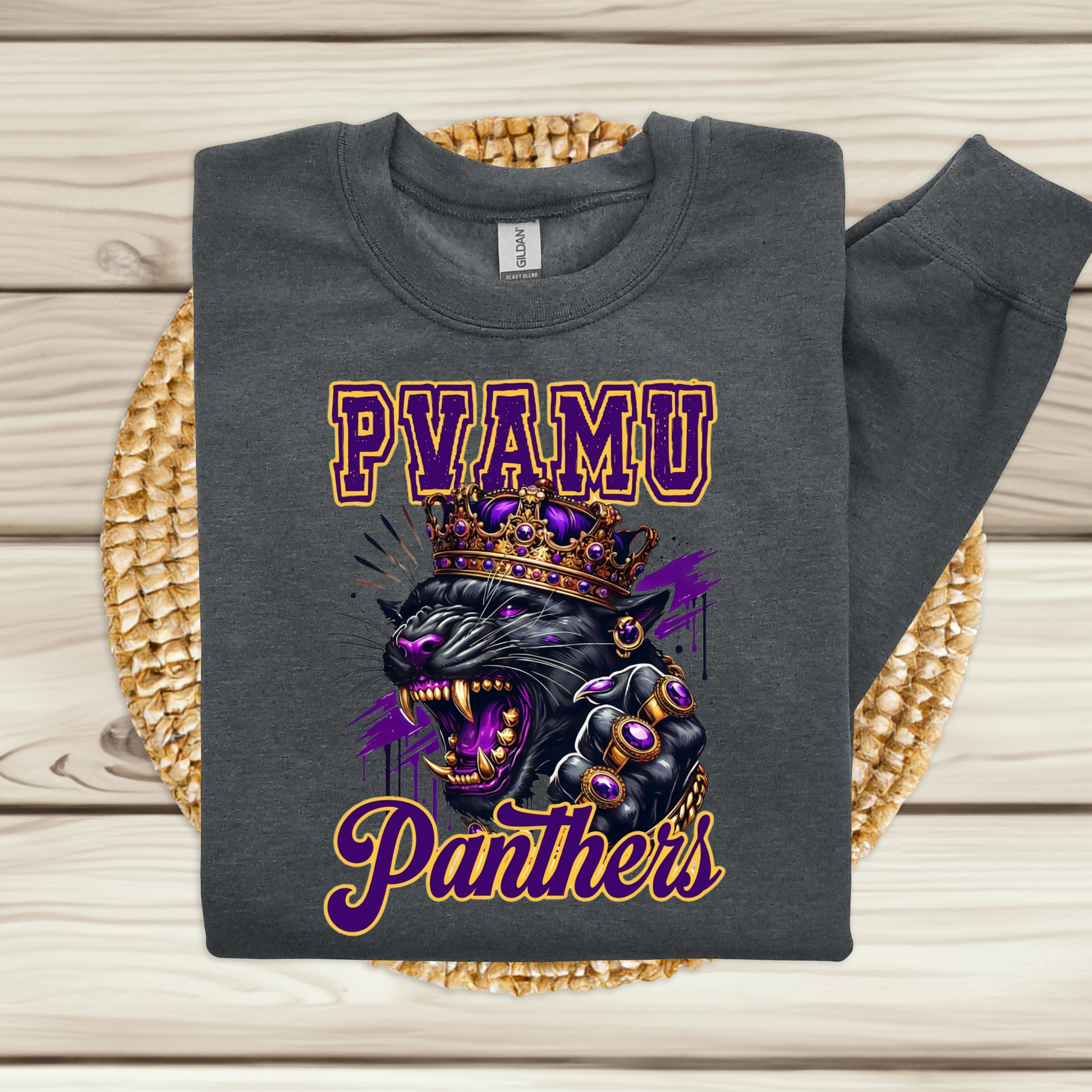 A dark gray sweatshirt laid flat on a woven round mat over a light wood plank surface. The sweatshirt has a large front graphic with the text 'PVAMU Panthers' in purple and gold, plus a crowned panther illustration in black, purple, and gold.