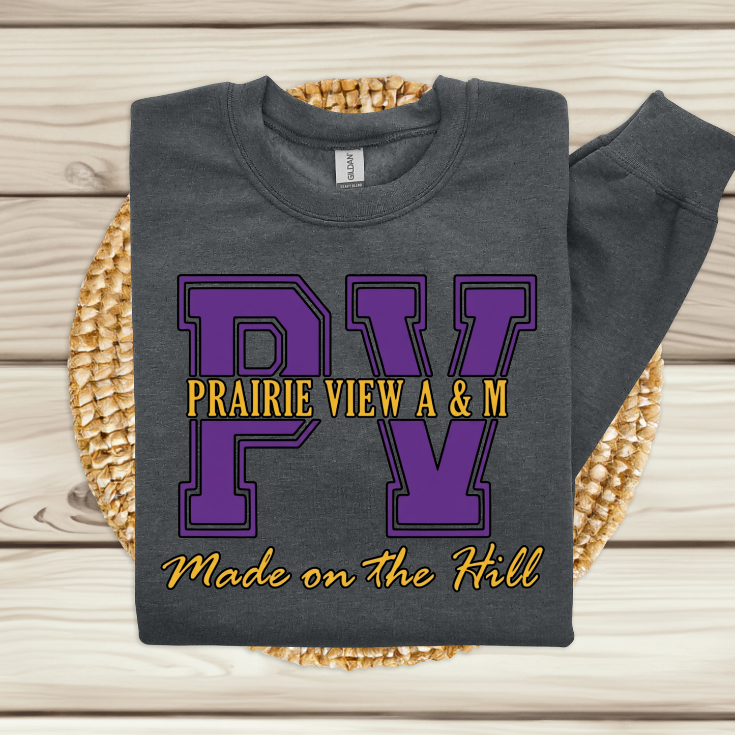 A dark gray sweatshirt laid flat on a woven mat over a light wood plank background. The sweatshirt has a large purple 'PV' graphic with yellow text reading 'PRAIRIE VIEW A & M' and 'Made on the Hill' in yellow script below.