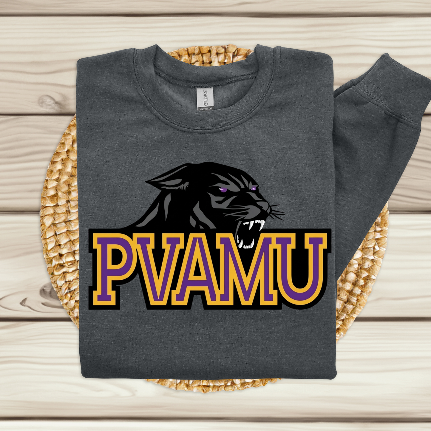 A dark gray sweatshirt laid flat on a woven round mat over a light wood plank surface. The sweatshirt has a large graphic of a black panther head with purple eyes above the text 'PVAMU' in purple and gold outlined letters.