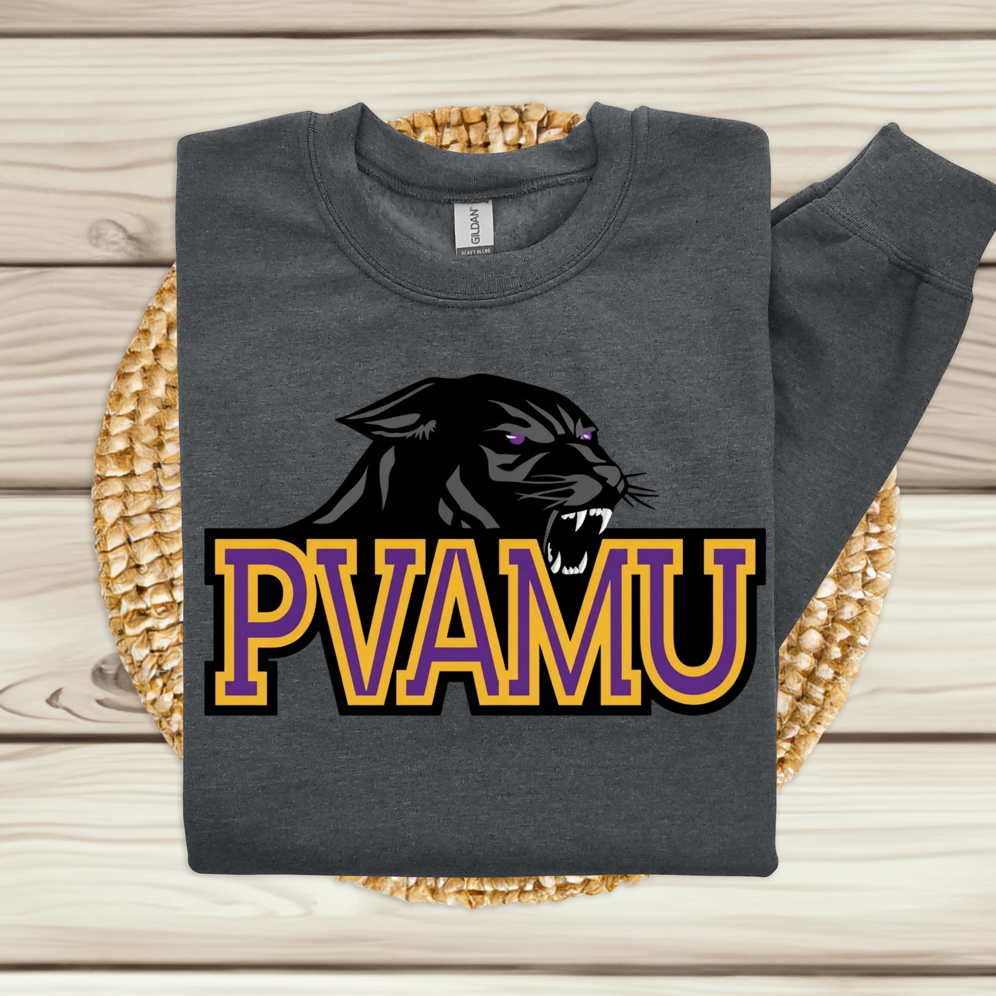 A dark gray sweatshirt laid flat on a woven round mat over a light wood plank surface. The sweatshirt has a large graphic of a black panther head with purple eyes above the text 'PVAMU' in purple and gold outlined letters.
