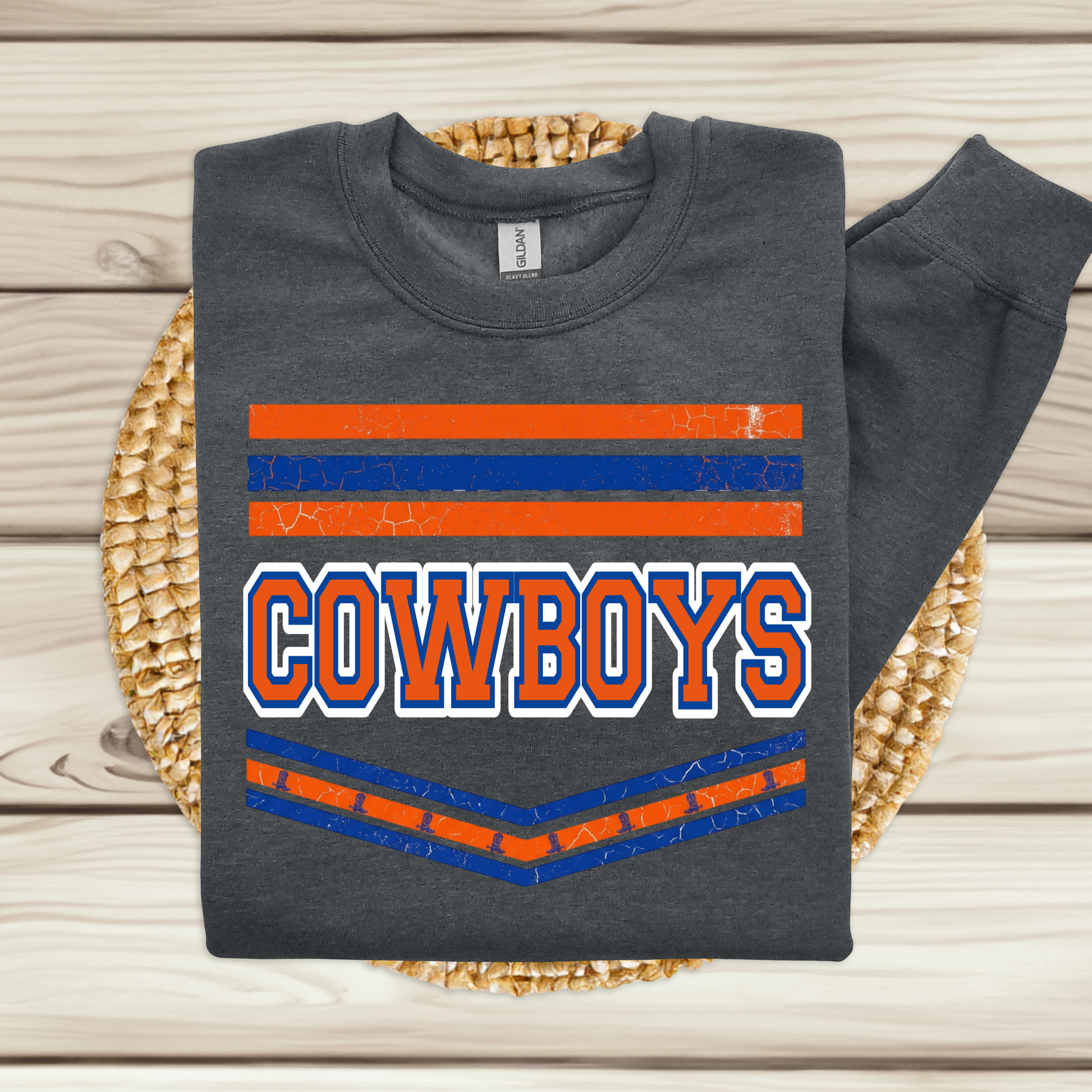 A dark gray long-sleeve crewneck sweatshirt with a distressed Cowboys graphic, displayed flat on a woven straw mat over a light wood plank background.