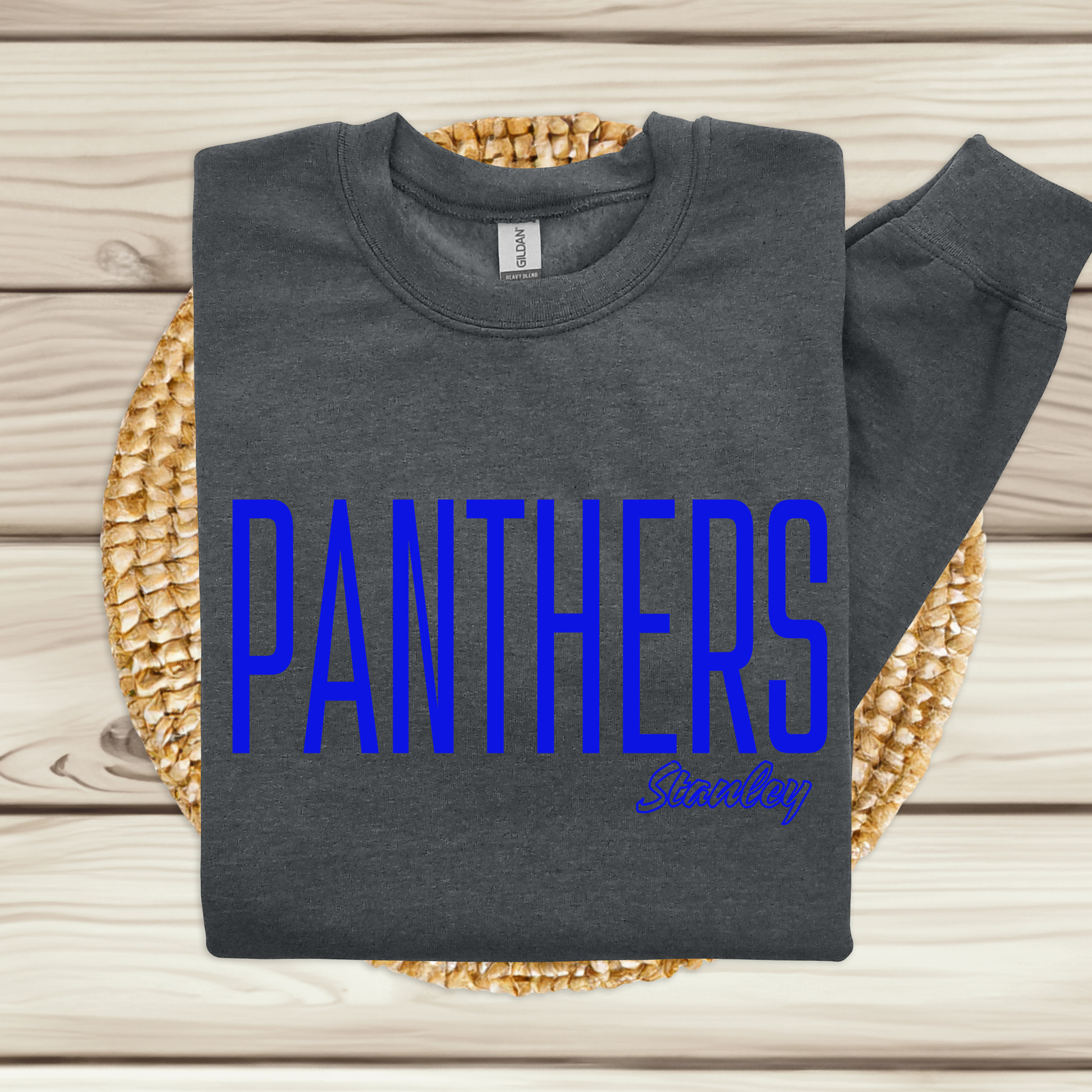 A dark gray sweatshirt with 'PANTHERS' printed in bold blue letters and 'Sunday' in cursive, displayed on a woven mat over a light wood plank background.