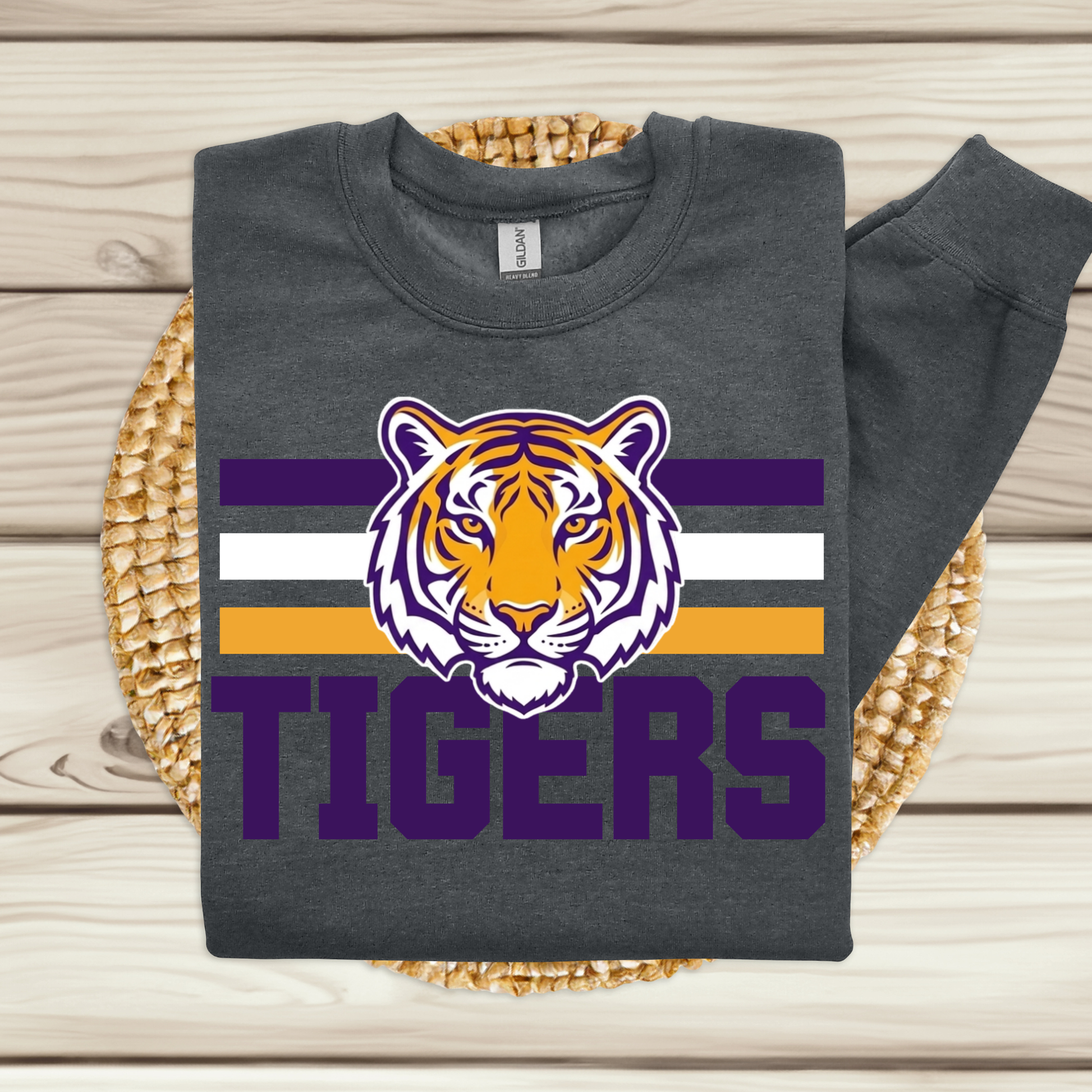 A dark gray sweatshirt laid flat on a woven round mat over a light wood plank surface. The sweatshirt has a large tiger head graphic above the word "TIGERS" with purple, white, and orange stripes behind it.