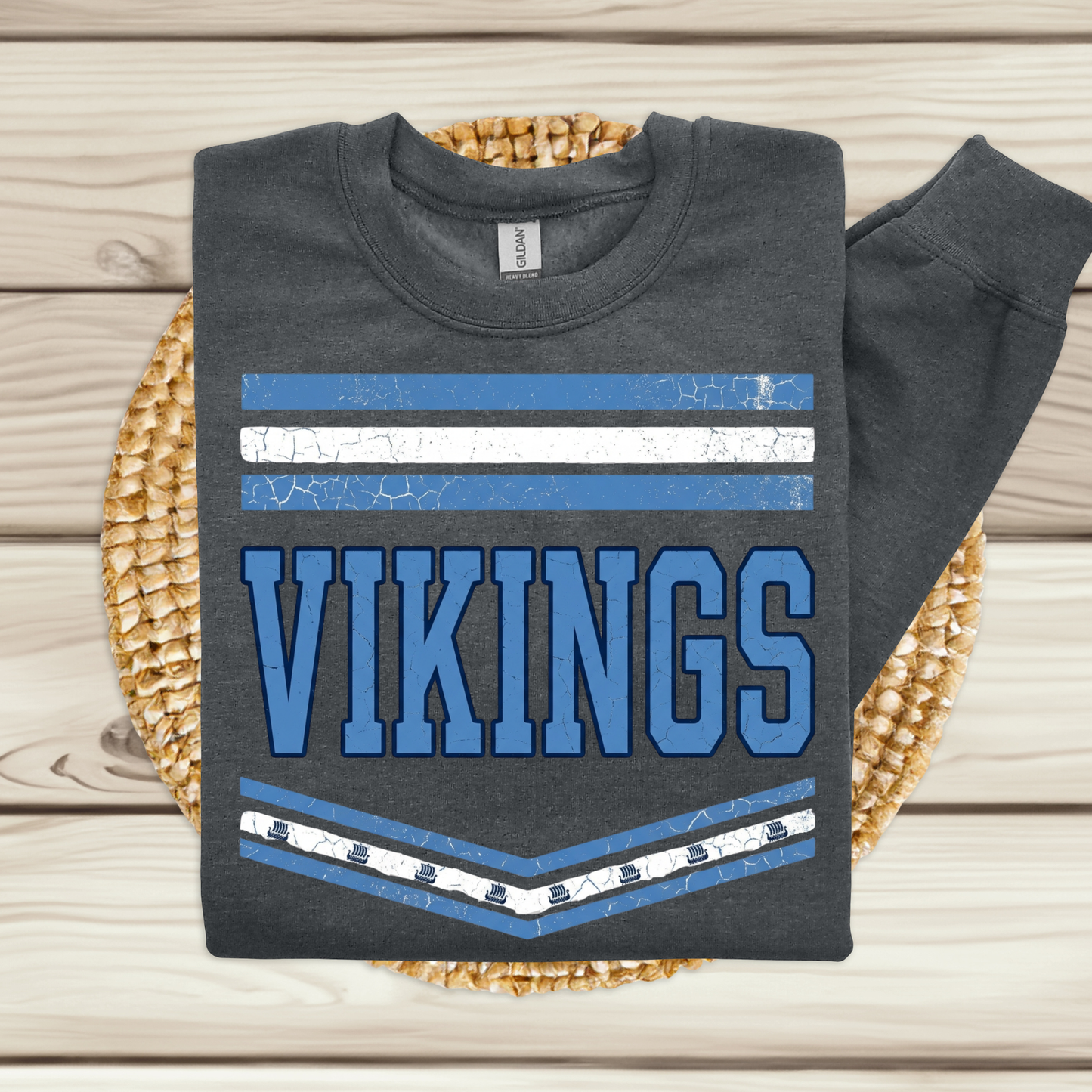 A dark gray long-sleeve t-shirt with a blue and white Vikings graphic, folded and displayed on a woven straw mat over a light wood plank background.