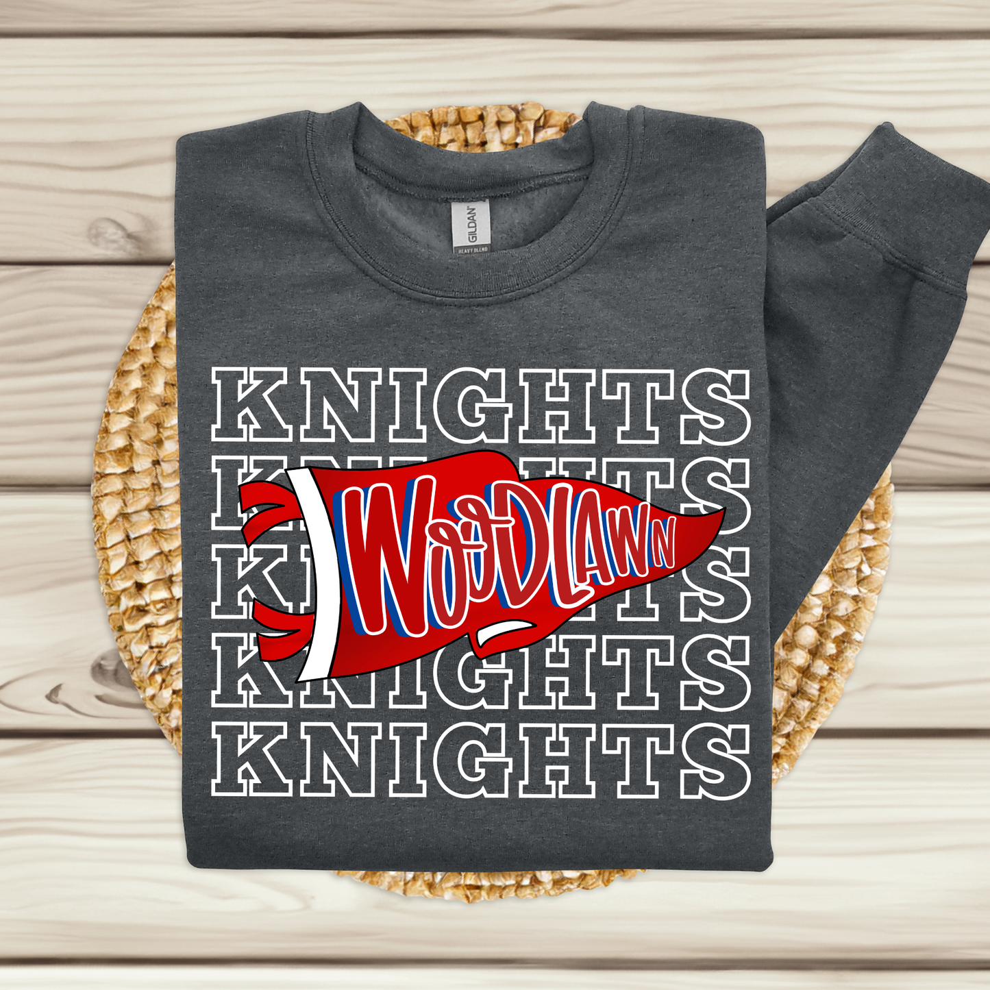 A dark charcoal gray sweatshirt laid flat on a woven round mat over a light wood plank surface. The sweatshirt has a large front graphic with repeated white outlined 'KNIGHTS' text and a red pennant shape across the chest reading 'Woodland' in white and blue.