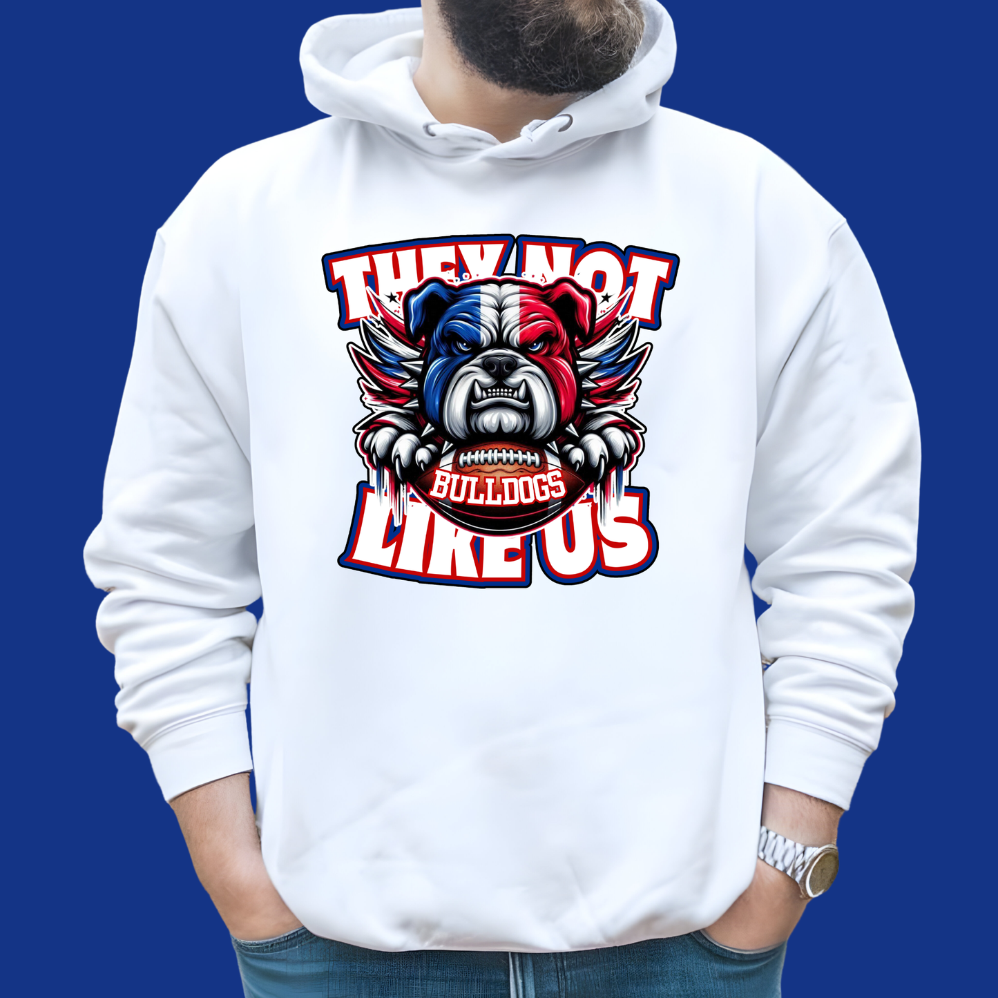 A white pullover hoodie with a large graphic print of a bulldog wearing a football helmet and the words 'THEY NOT LIKE US' and 'BULLDOGS' on the front, shown on a model against a solid blue background.