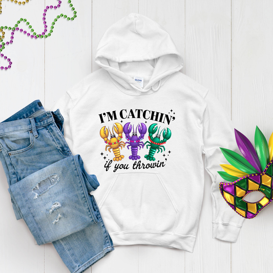 White hoodie with colorful lobster design and text, paired with denim shorts and a Mardi Gras mask on a light wooden surface.