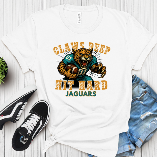 White t-shirt with a leopard graphic and text on a wooden surface with shoes and jeans. Jaguars Mascot Shirt – Fierce Jaguars "Claws Deep Hit Hard" Spirit Tee