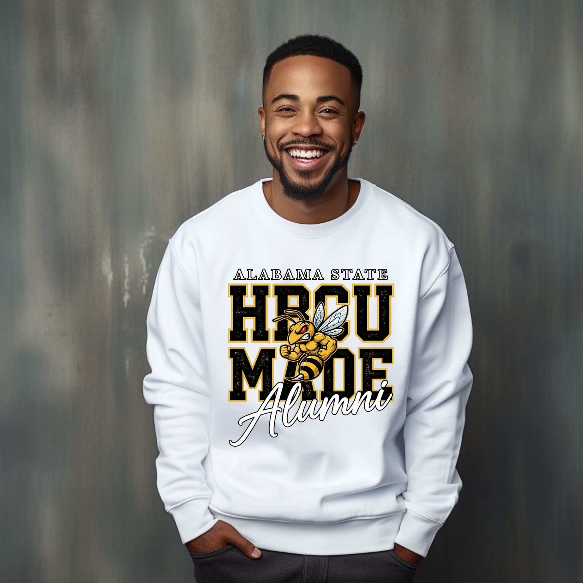 A smiling man wearing a white crewneck sweatshirt with a graphic that reads 'ALABAMA STATE HBCU MADE Alumni' with a bee illustration, standing against a textured gray background.