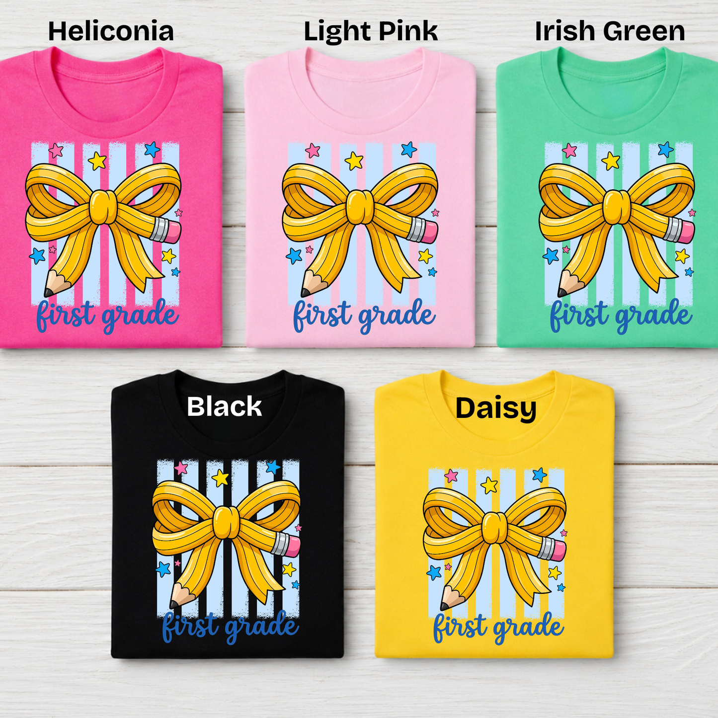 Five folded graphic T-shirts arranged in a grid on a light wood background, labeled Heliconia, Light Pink, Irish Green, Black, and Daisy. Each shirt shows a large yellow ribbon bow with pencils on a white and pastel striped panel above the words 'first grade'.