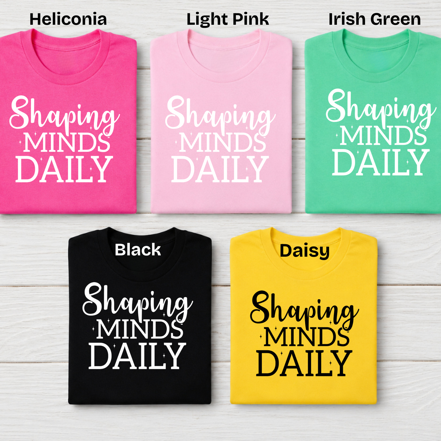 Five folded graphic T-shirts arranged in a 2x3 grid on a light wood background, labeled with color names at the top: Heliconia, Light Pink, Irish Green, Black, and Daisy. Each shirt has the phrase 'Shaping MINDS DAILY' printed in white or black script and block lettering.