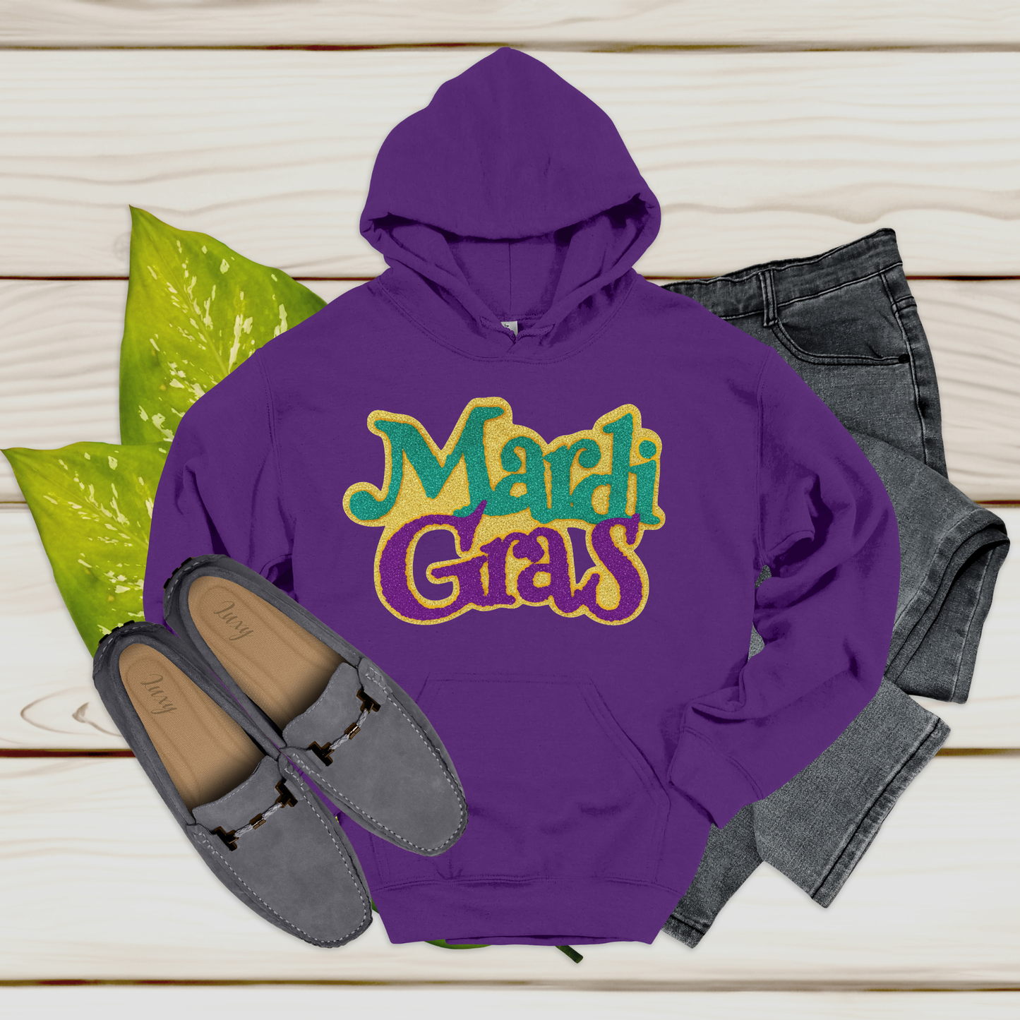 Purple hoodie with 'Mardi Gras' text, gray loafers, and jeans on a wooden surface