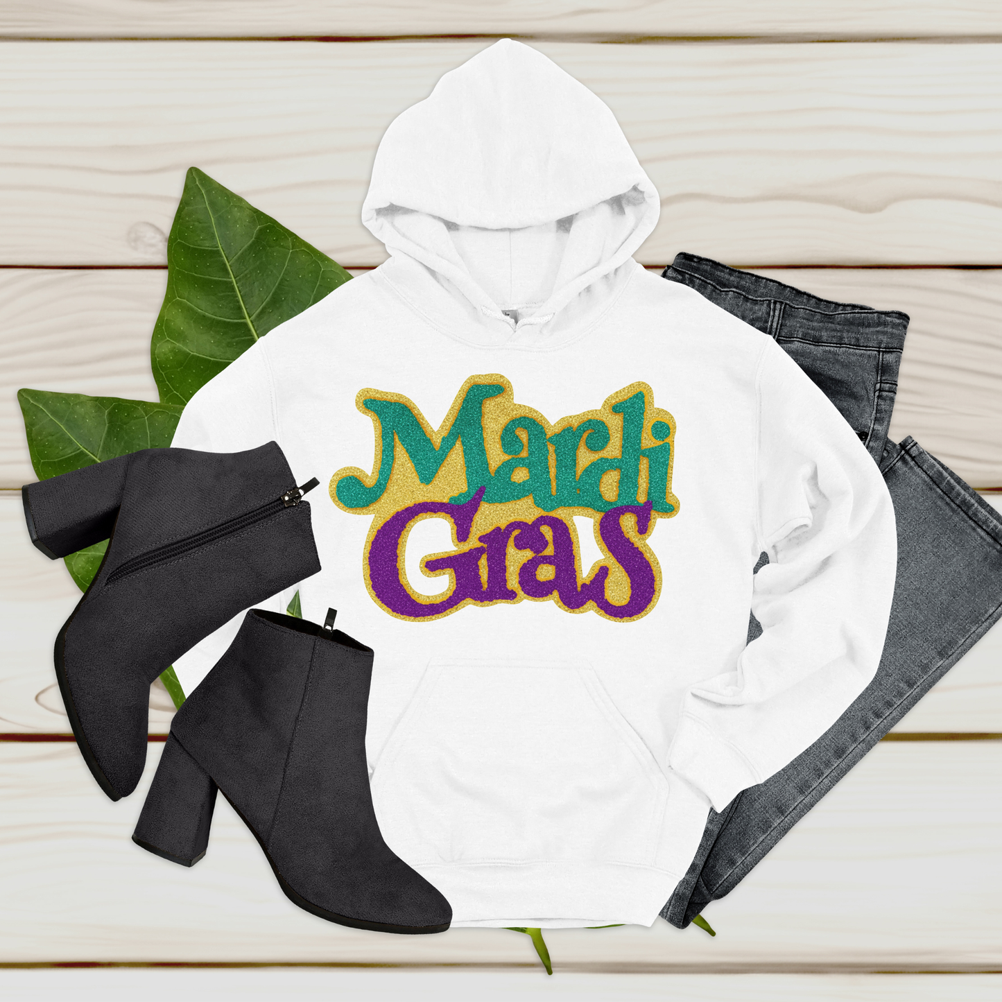 White hoodie with 'Mardi Gras' text, black ankle boots, and gray jeans on a wooden surface.