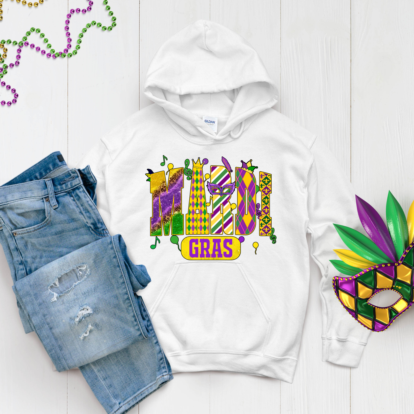 White hoodie with Mardi Gras design, denim shorts, and a mask on a light wooden surface.