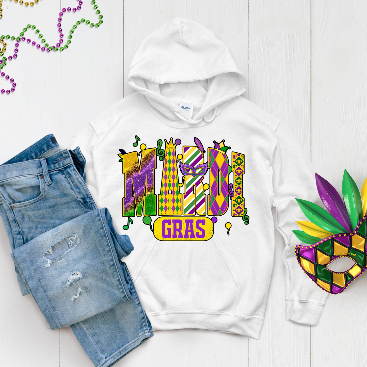 White hoodie with Mardi Gras design, blue jeans, and a mask on a light wooden surface.