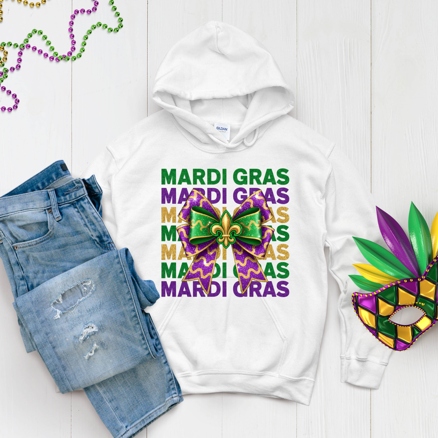 White hoodie with Mardi Gras-themed design, paired with denim shorts and a mask on a light wooden surface.