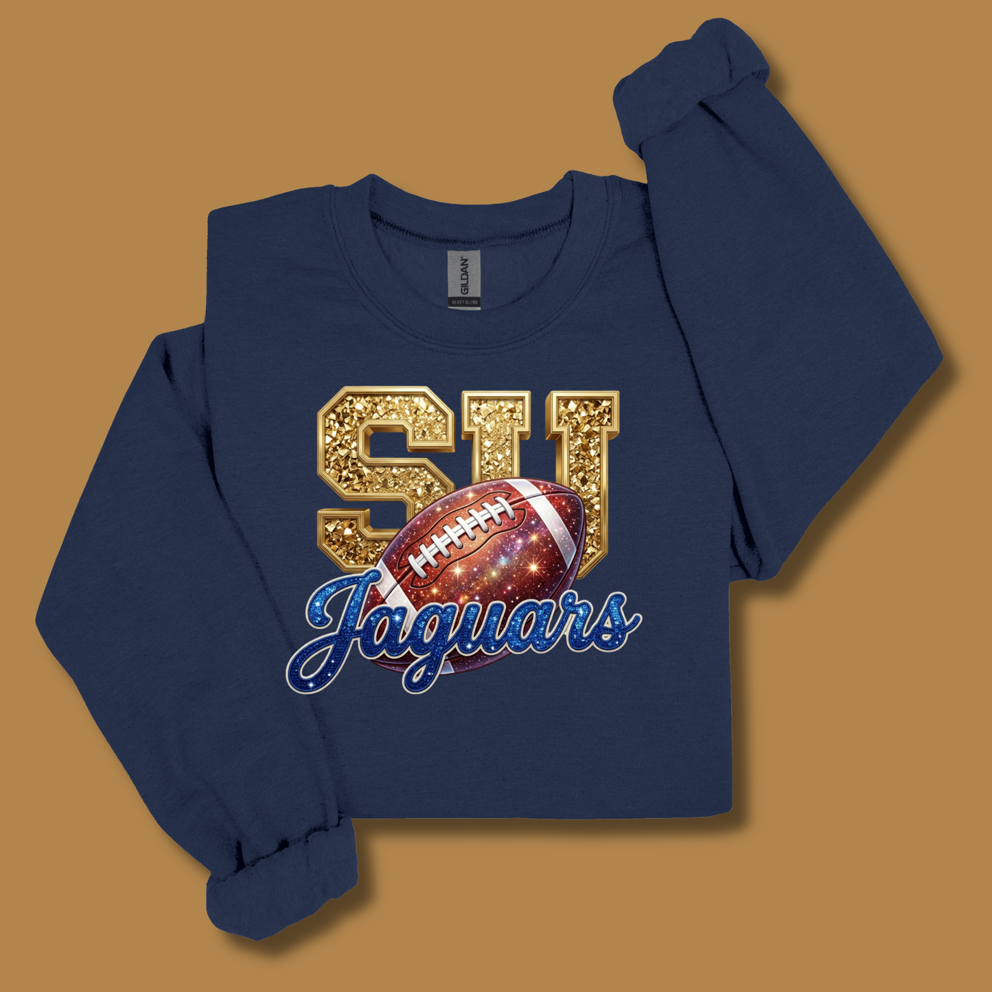 A navy blue long-sleeve sweatshirt with a graphic print featuring gold glittery 'SU' letters, a football, and 'Jaguars' in blue script, displayed on a solid tan background.