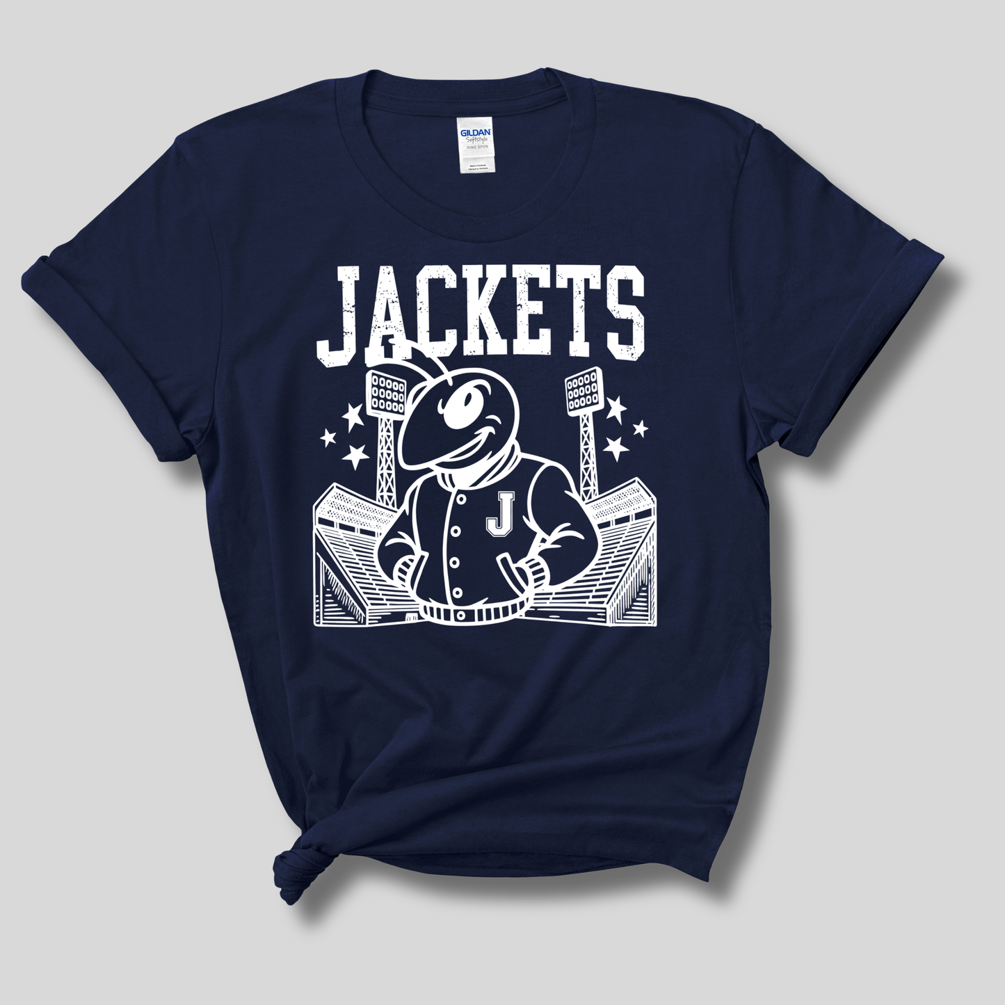A navy blue short-sleeve t-shirt with a white graphic print featuring the word 'JACKETS' and a cartoon mascot in a jacket standing in front of stadium lights. The shirt is displayed flat against a neutral gray background.