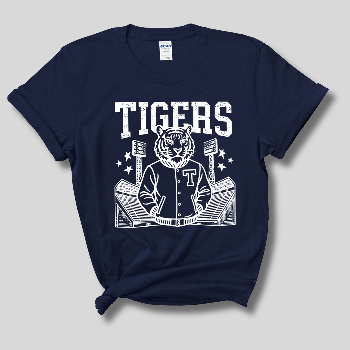 A navy blue short-sleeve t-shirt with a white graphic print of a tiger mascot wearing a baseball jersey, standing in front of a stadium, with the word 'TIGERS' printed above. The shirt is shown flat against a neutral gray background.