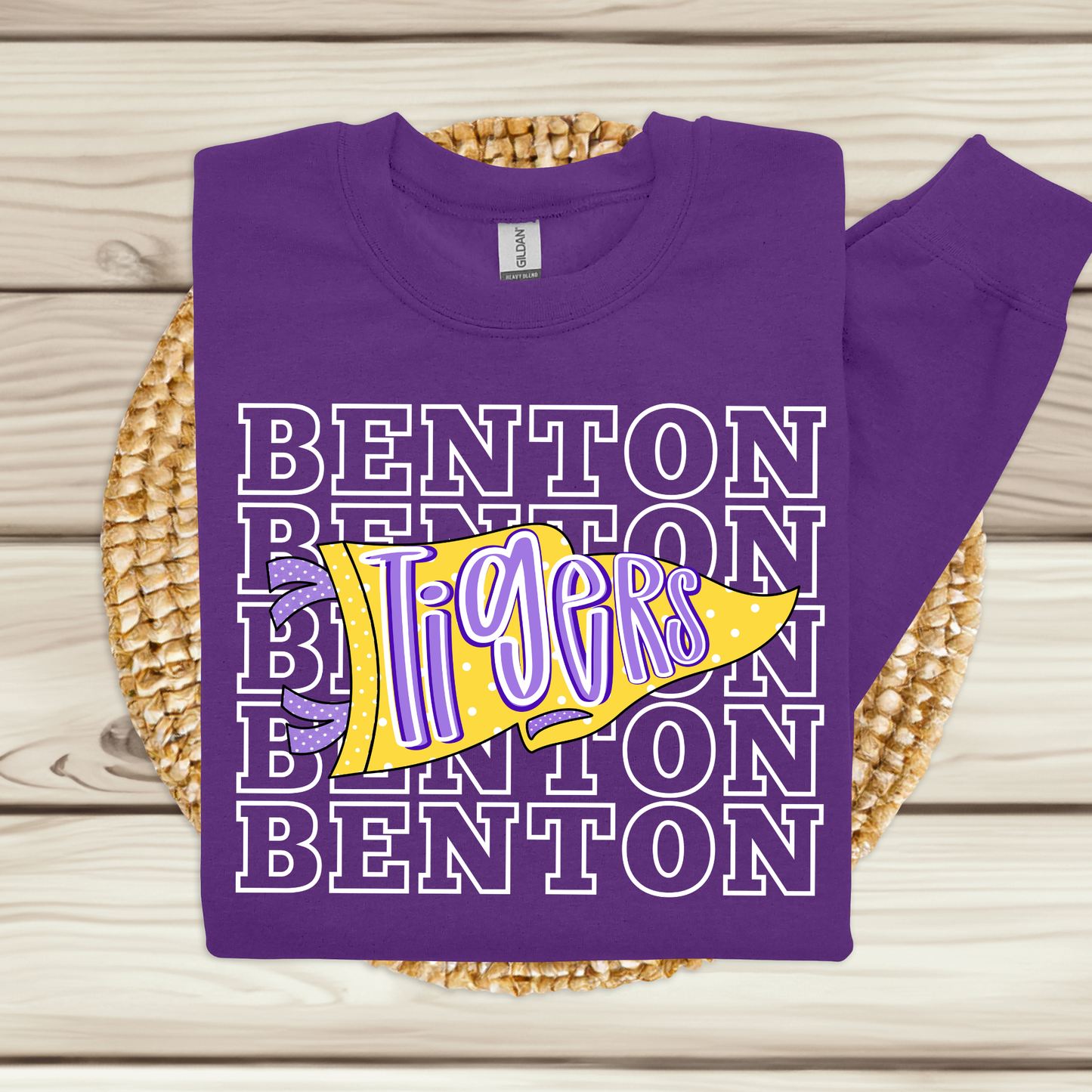 A purple sweatshirt laid flat on a woven round mat over light wood planks. The sweatshirt has a large front graphic with repeated white outlined 'BENTON' text and a yellow pennant shape reading 'Tigers' in purple with small yellow stars.