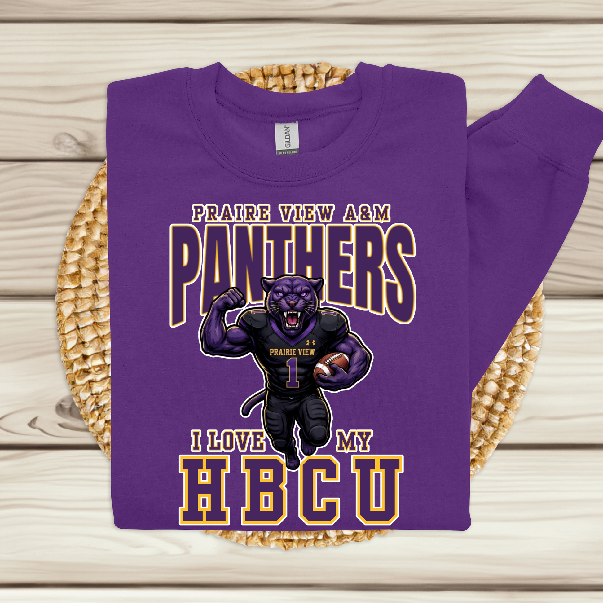 A purple crewneck sweatshirt with a 'Prairie View 1876 A & M University' graphic in gold and white, displayed flat on a woven round mat over a light wood plank background.
