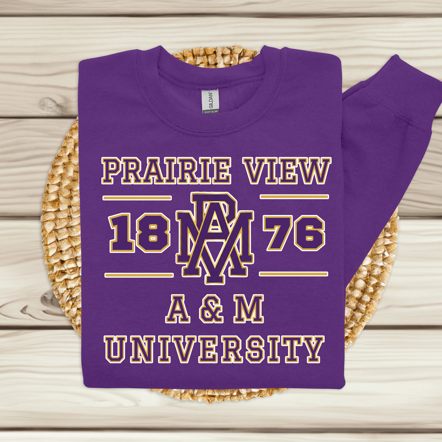 A purple crewneck sweatshirt with a 'Prairie View 1876 A & M University' graphic in gold and white, displayed flat on a woven round mat over a light wood plank background.