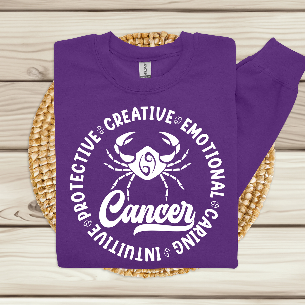 A purple crewneck sweatshirt with a white graphic print of a Cancer zodiac symbol and the word 'Cancer' surrounded by adjectives, laid flat on a woven placemat over a light wood background.