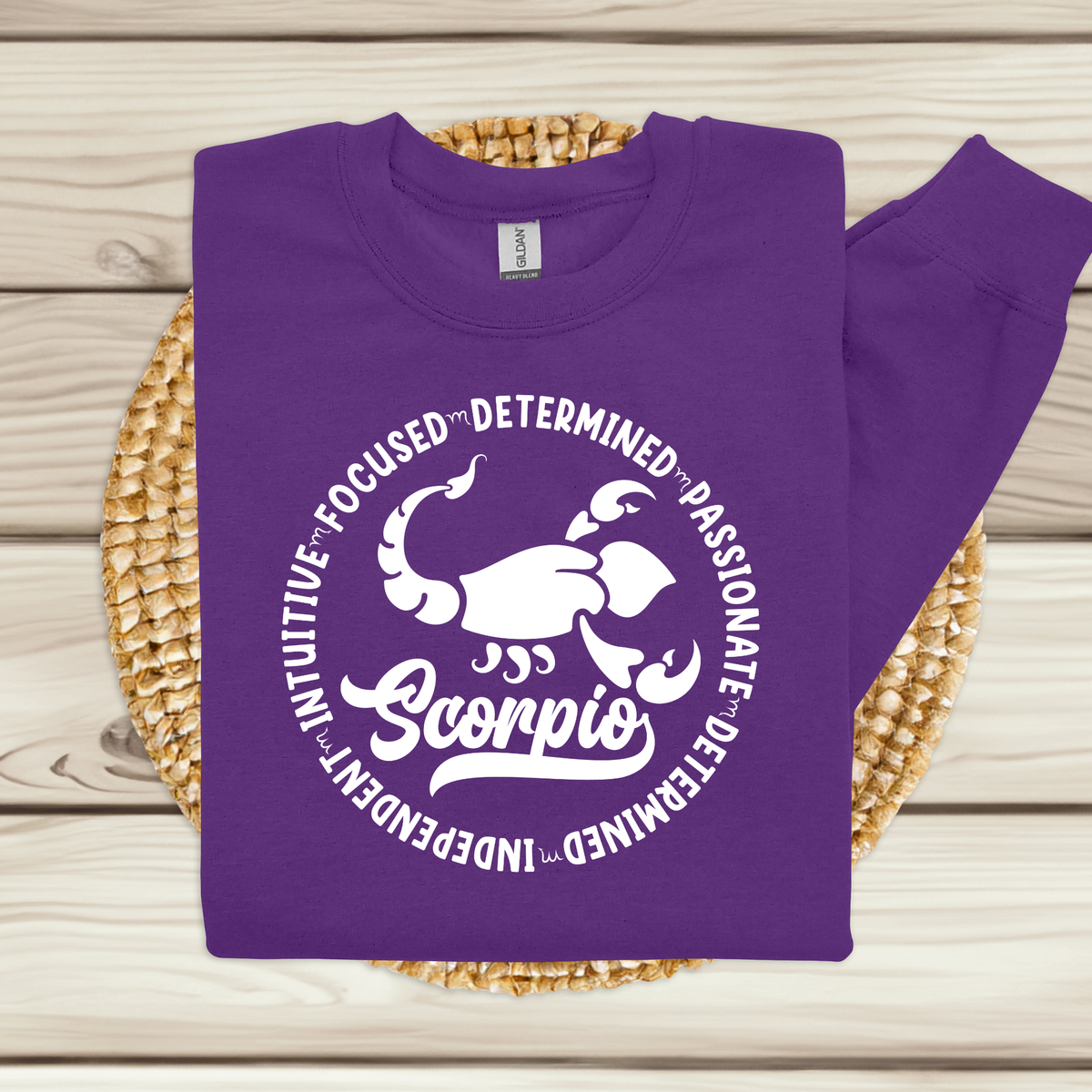 Woman wearing a purple crewneck sweatshirt featuring a white circular Scorpio zodiac graphic with a scorpion illustration, the word "Scorpio," and trait keywords like intuitive, focused, determined, passionate, and independent.