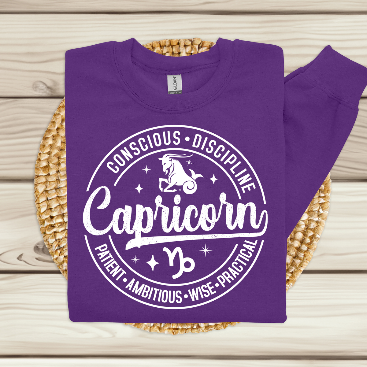 A purple long-sleeve sweatshirt laid flat on a woven round mat over light wood planks. The sweatshirt has a large white circular graphic with the word 'Capricorn,' a zodiac symbol, stars, and the words 'Conscious • Discipline,' 'Patient • Ambitious • Wise • Practical.
