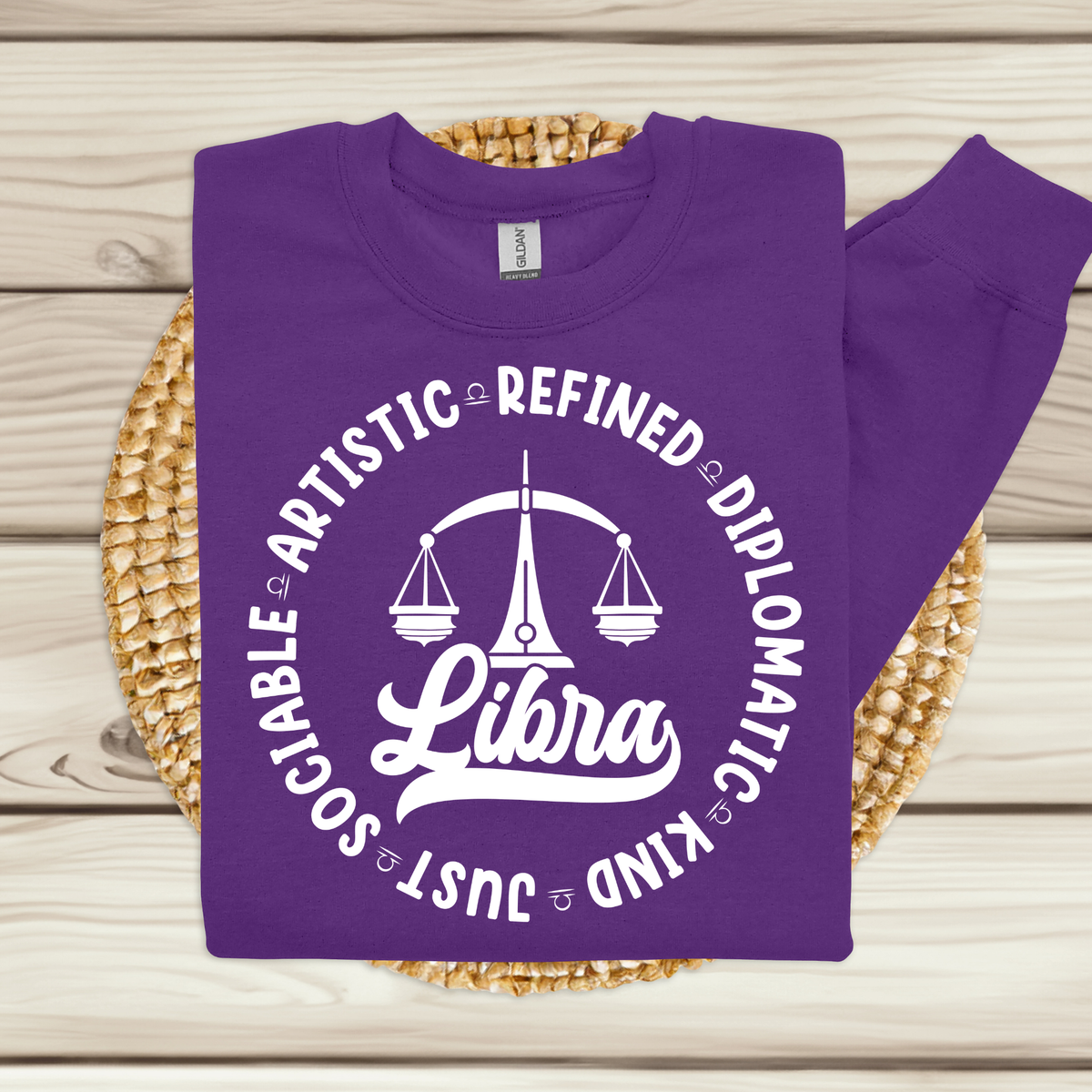 A purple crewneck sweatshirt with a white graphic print featuring a Libra zodiac symbol and the words 'Libra' and 'Just Sociable Artistic Refined Diplomatic Kind', displayed on a woven mat over a light wood background.