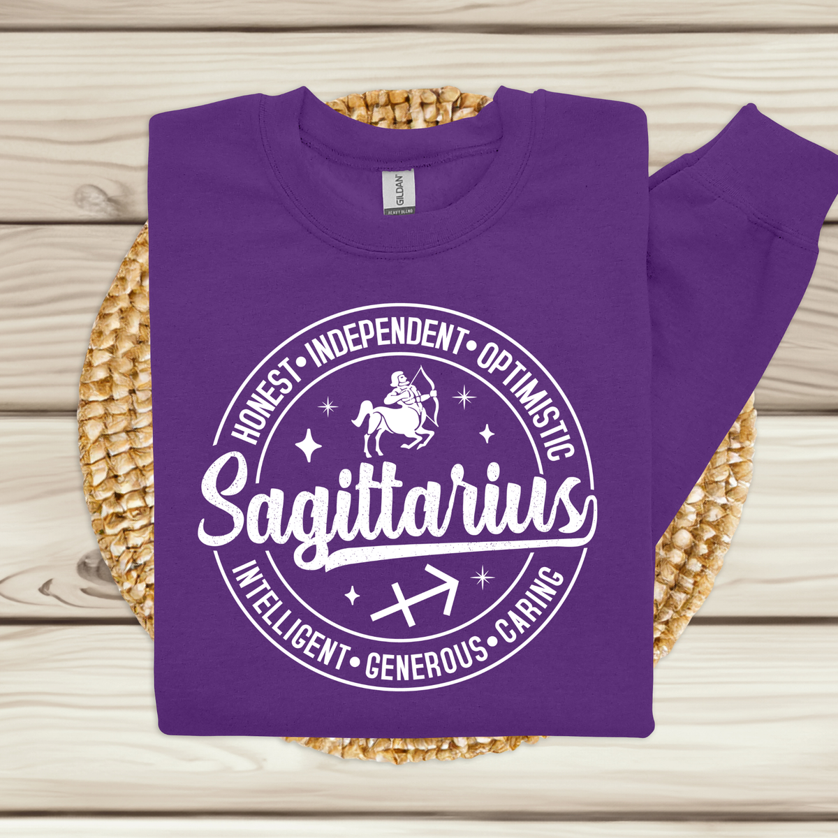 A purple long-sleeve sweatshirt with a white circular graphic reading 'Sagittarius' and listing traits like Honest, Independent, Optimistic, Intelligent, Generous, and Caring. The sweatshirt is laid flat on a woven round mat over a light wood plank background.