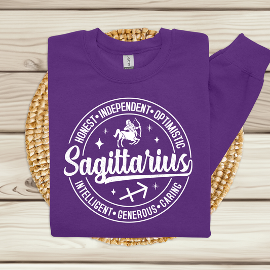 A purple long-sleeve sweatshirt with a white circular graphic reading 'Sagittarius' and listing traits like Honest, Independent, Optimistic, Intelligent, Generous, and Caring. The sweatshirt is laid flat on a woven round mat over a light wood plank background.