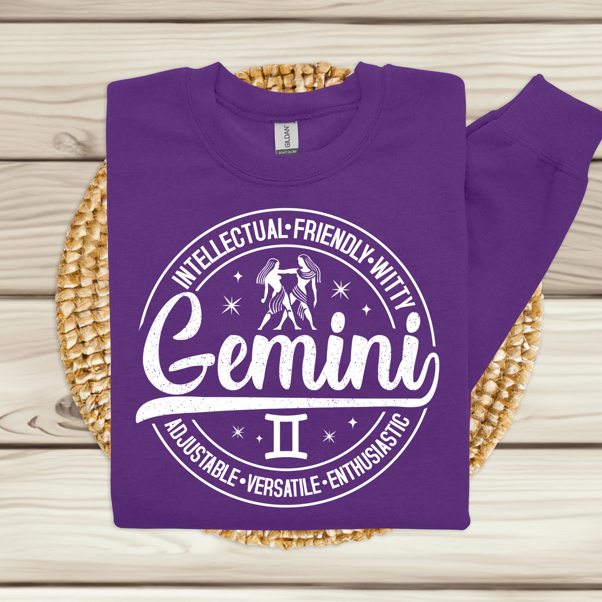 purple crewneck t-shirt laid flat, featuring a circular white Gemini zodiac graphic with the twins illustration, the word "Gemini," and trait keywords like intellectual, friendly, witty, enthusiastic, versatile, and adjustable.