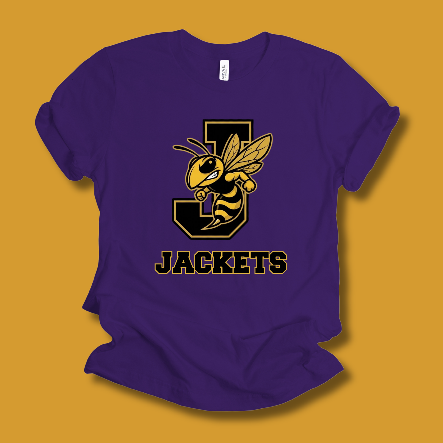 A purple short-sleeve t-shirt with a yellow and black wasp mascot graphic over a large letter 'J' and the word 'JACKETS' printed on the front, displayed on a solid mustard yellow background.