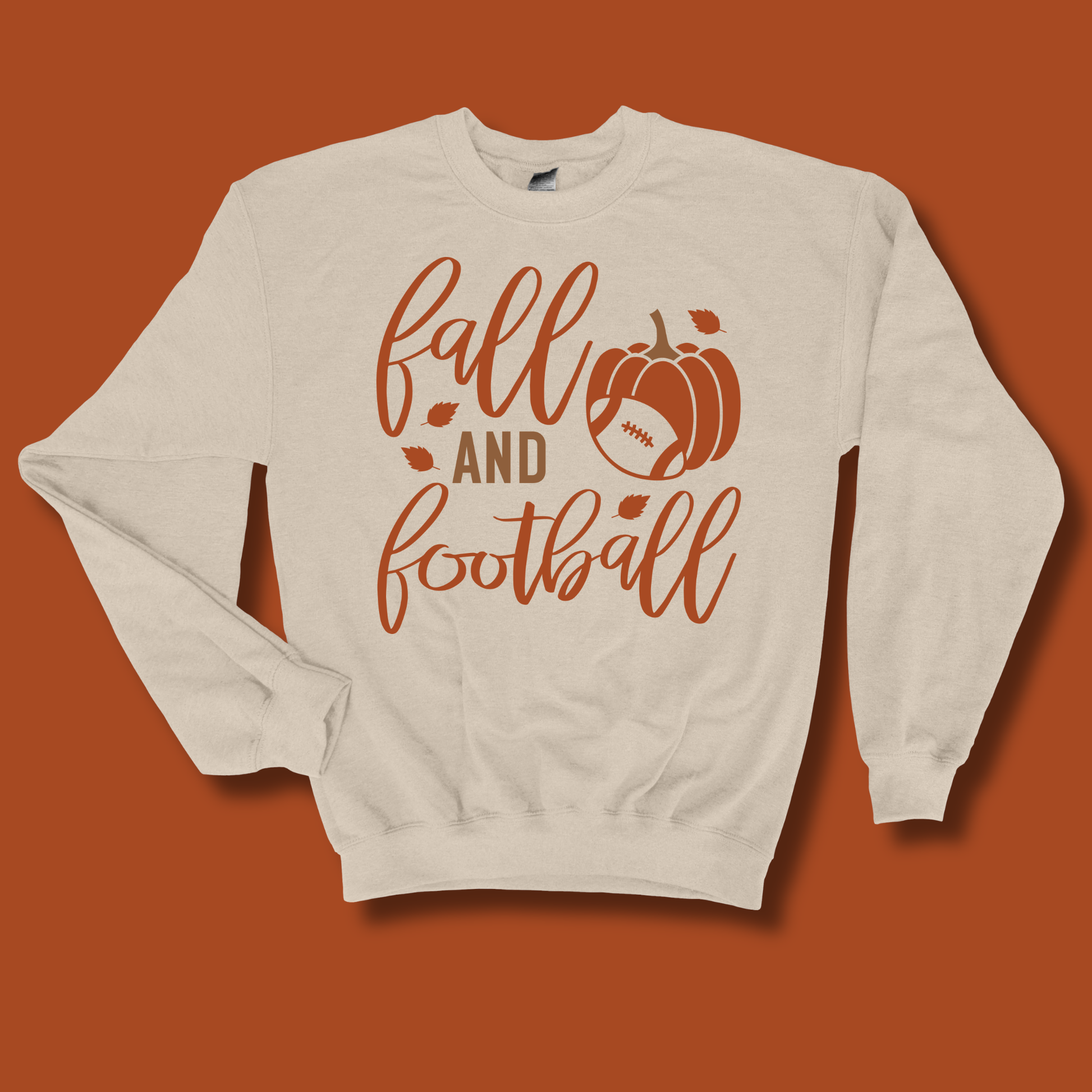 An off-white crewneck sweatshirt with a 'fall and football' graphic in rust orange, featuring a pumpkin and football design, displayed against a solid rust orange background.