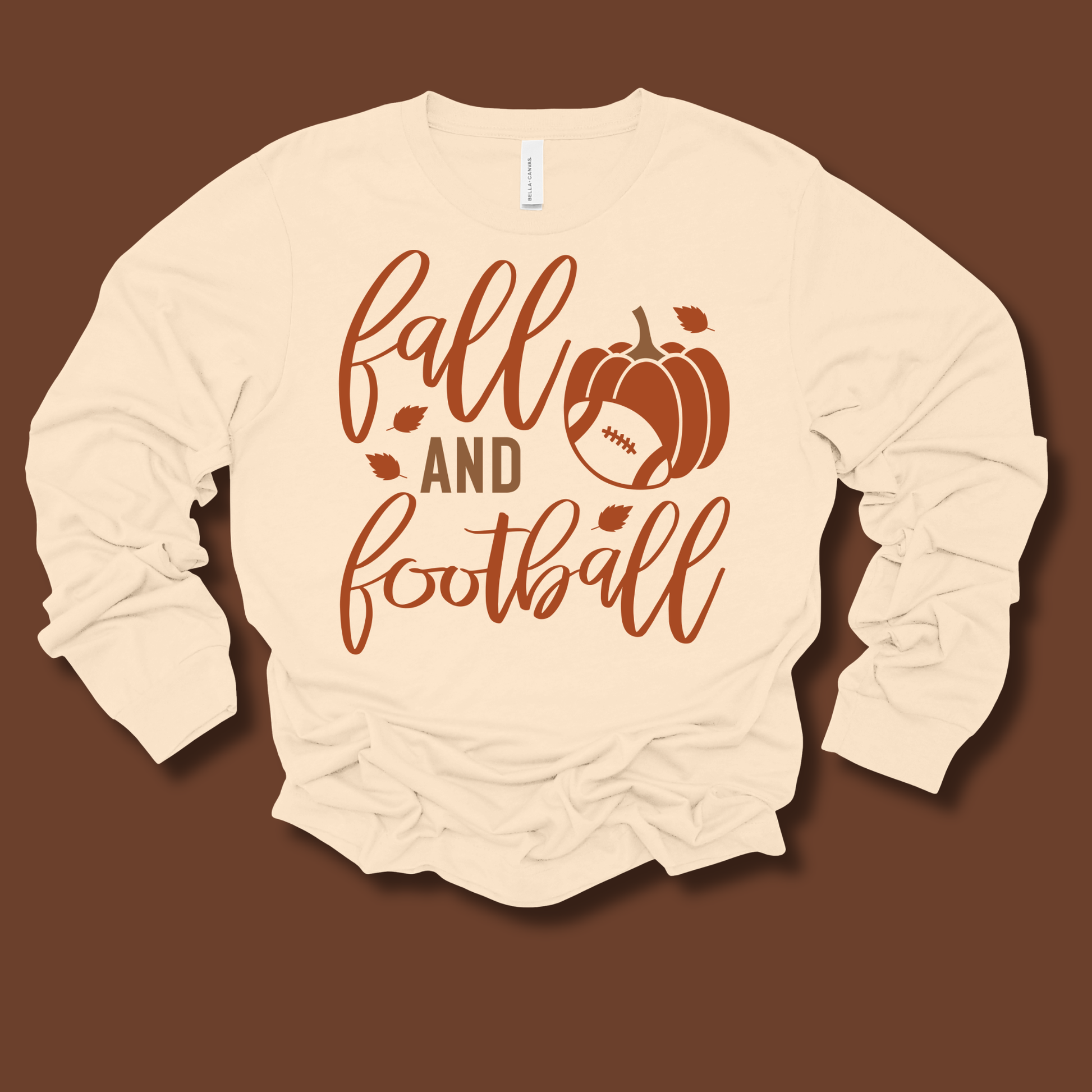 A cream-colored long-sleeve t-shirt with a 'fall and football' graphic print in brown, featuring a pumpkin and football design, laid flat against a solid brown background.
