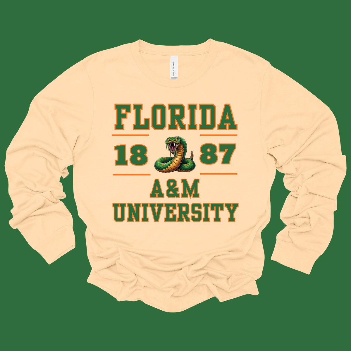 A cream-colored long-sleeve t-shirt with a green and orange graphic print reading 'FLORIDA 1887 A&M UNIVERSITY' with a snake illustration, displayed against a solid green background.