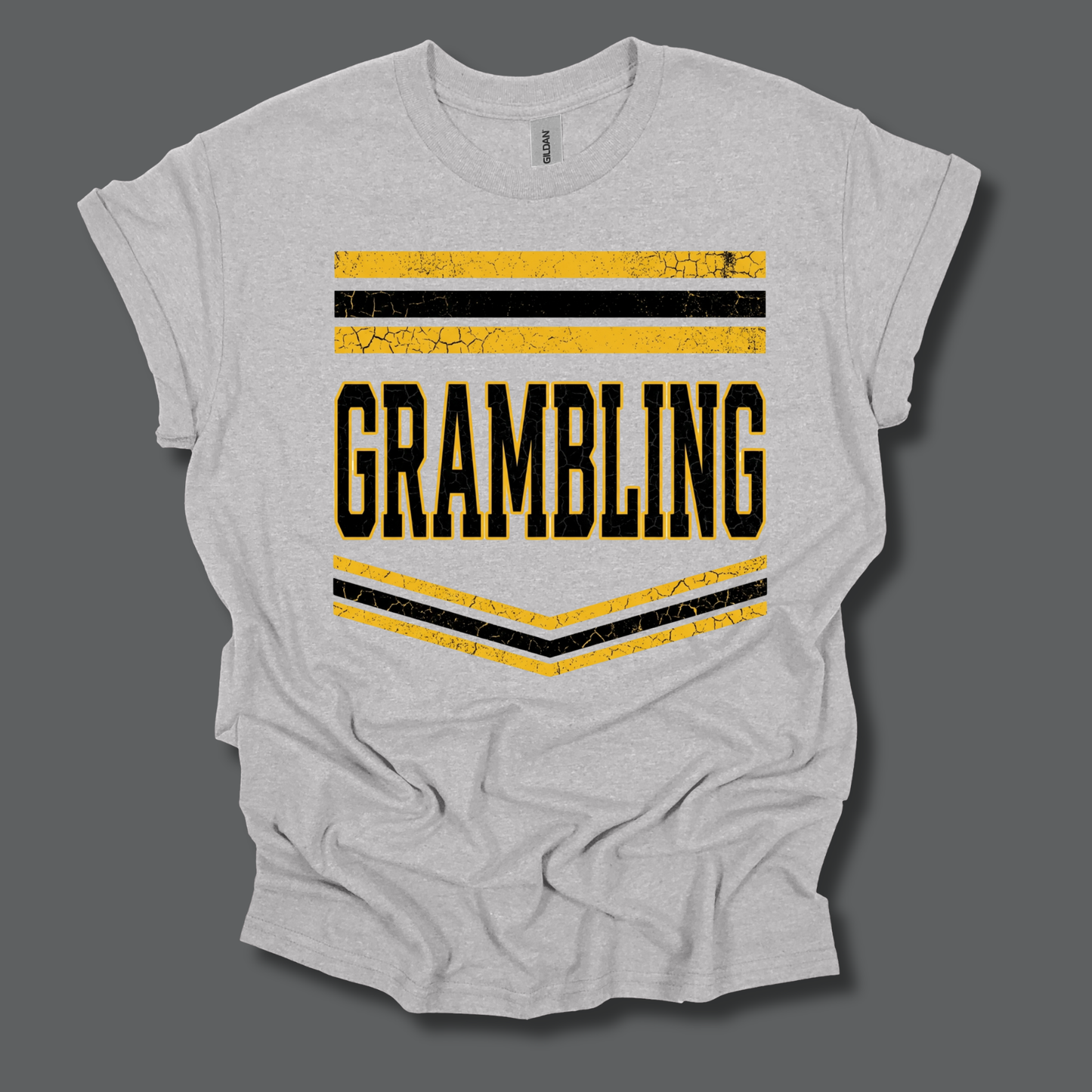 A light heather gray t-shirt with a distressed yellow and black graphic on the front that reads 'GRAMBLING' with horizontal stripes above and a V-shaped chevron below, displayed on a solid dark gray background.